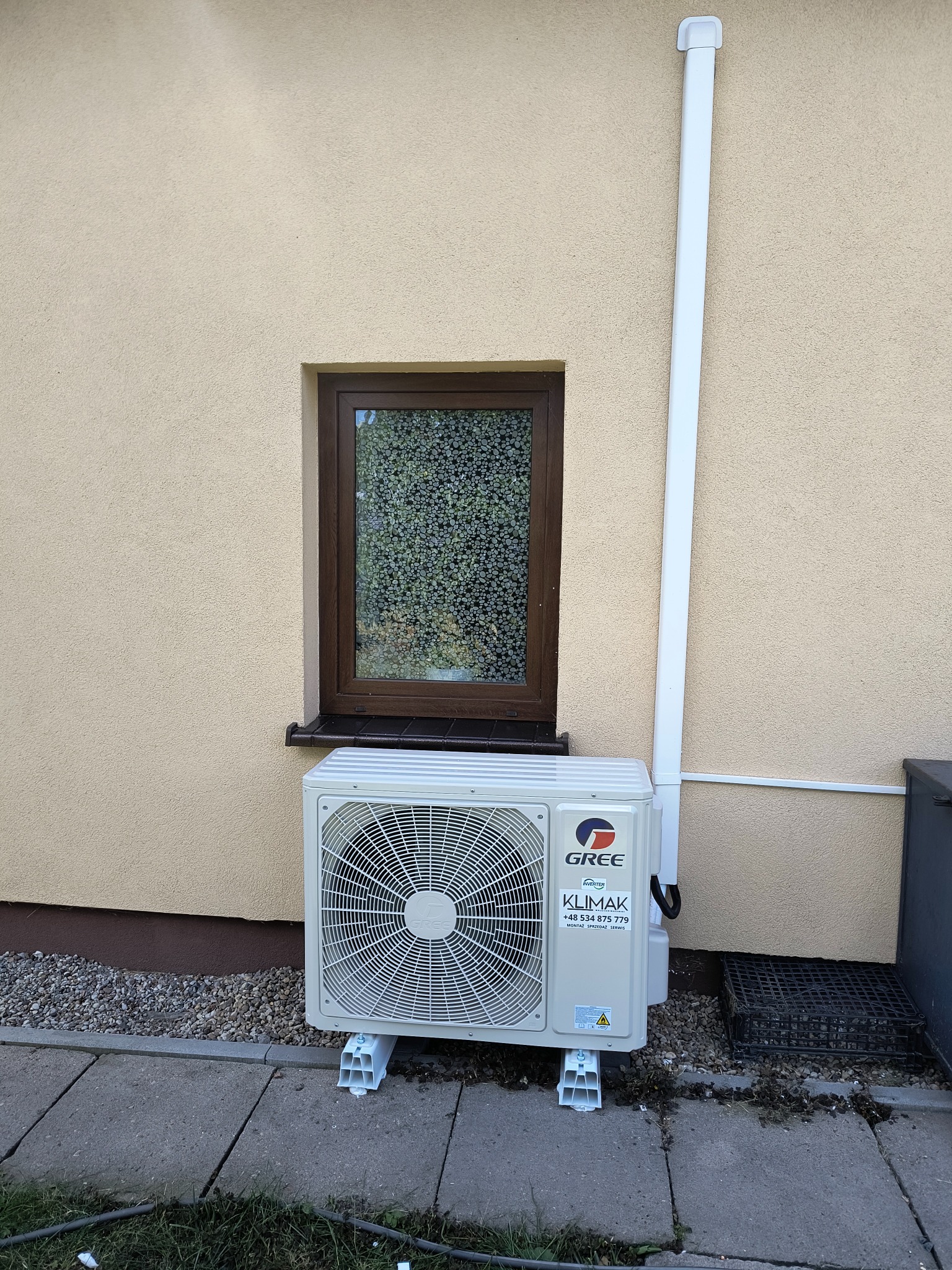 Zamontowana jednostka zewnętrzna klimatyzacji Gree Klimak na elewacji domu z brązowym oknem. Widoczna biała rura odprowadzająca skropliny i kostka brukowa.