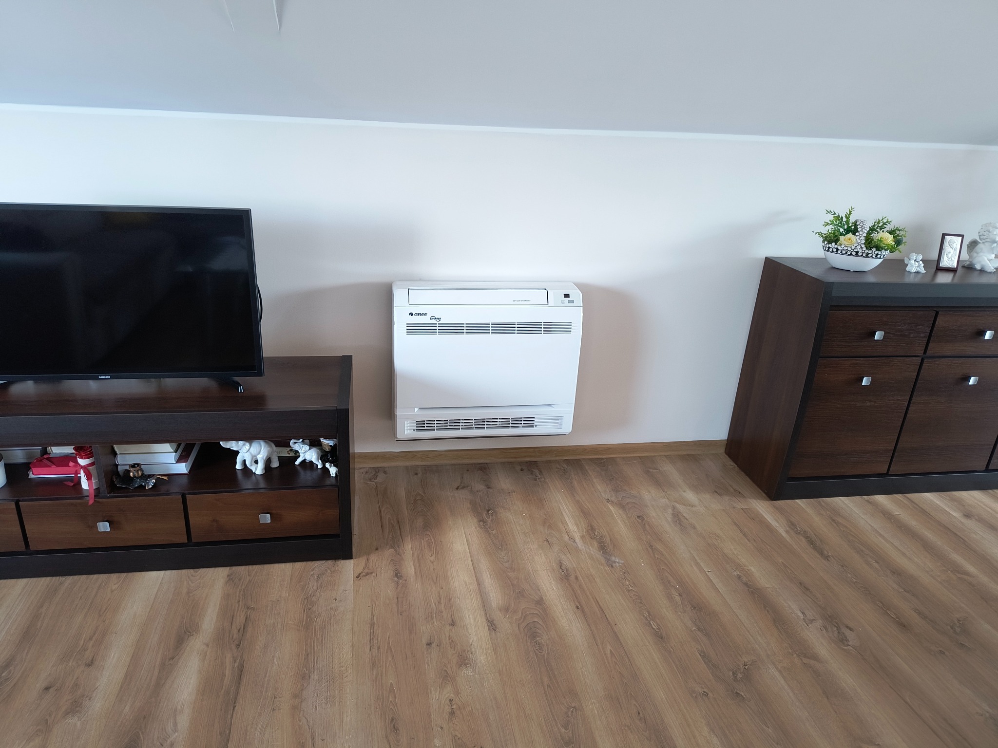 Biały klimatyzator podłogowy Gree Inverter w salonie, pomiędzy ciemnobrązową szafką RTV i komodą. Parkiet w odcieniach brązu. Ściana biała, skośna.
