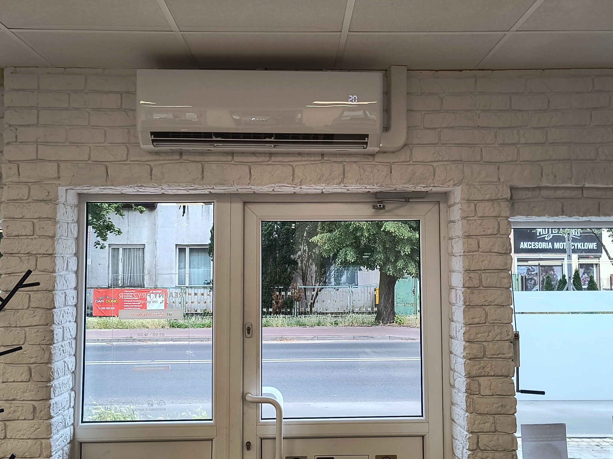 Biała klimatyzacja na ścianie z cegły nad przeszklonymi drzwiami wejściowymi. Widok na ulicę z zielenią i budynkami w tle.