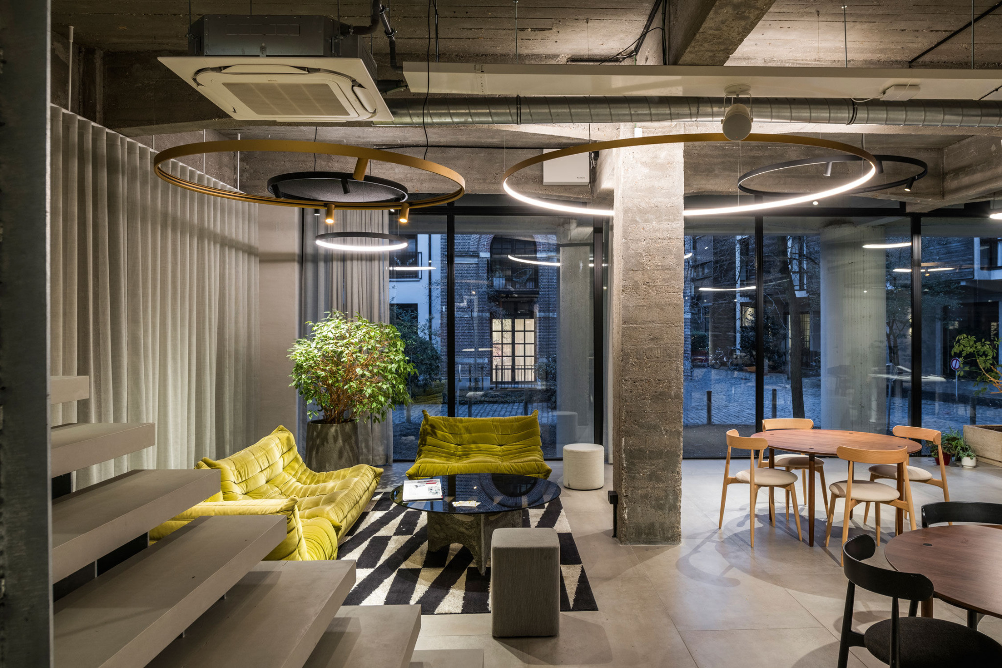 Nowoczesne wnętrze biurowe z betonowymi elementami, okrągłymi lampami i żółtymi sofami. Widoczne schody, stoliki i krzesła. Duże okna z widokiem na zewnątrz.