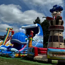 Kompleksowy, kolorowy plac zabaw, w tym dmuchany zamek w kształcie pirata na wieży, zjeżdżalnia w kształcie wieloryba i tęczowa zjeżdżalnia, ustawiony na trawie pod błękitnym niebem z chmurami.