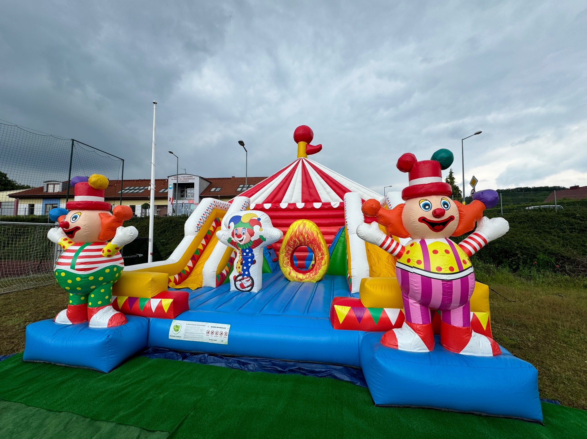 Kolorowy, dmuchany plac zabaw z motywem cyrkowym i dwoma dużymi figurami klaunów po bokach, ustawiony na zielonej trawie pod pochmurnym niebem.