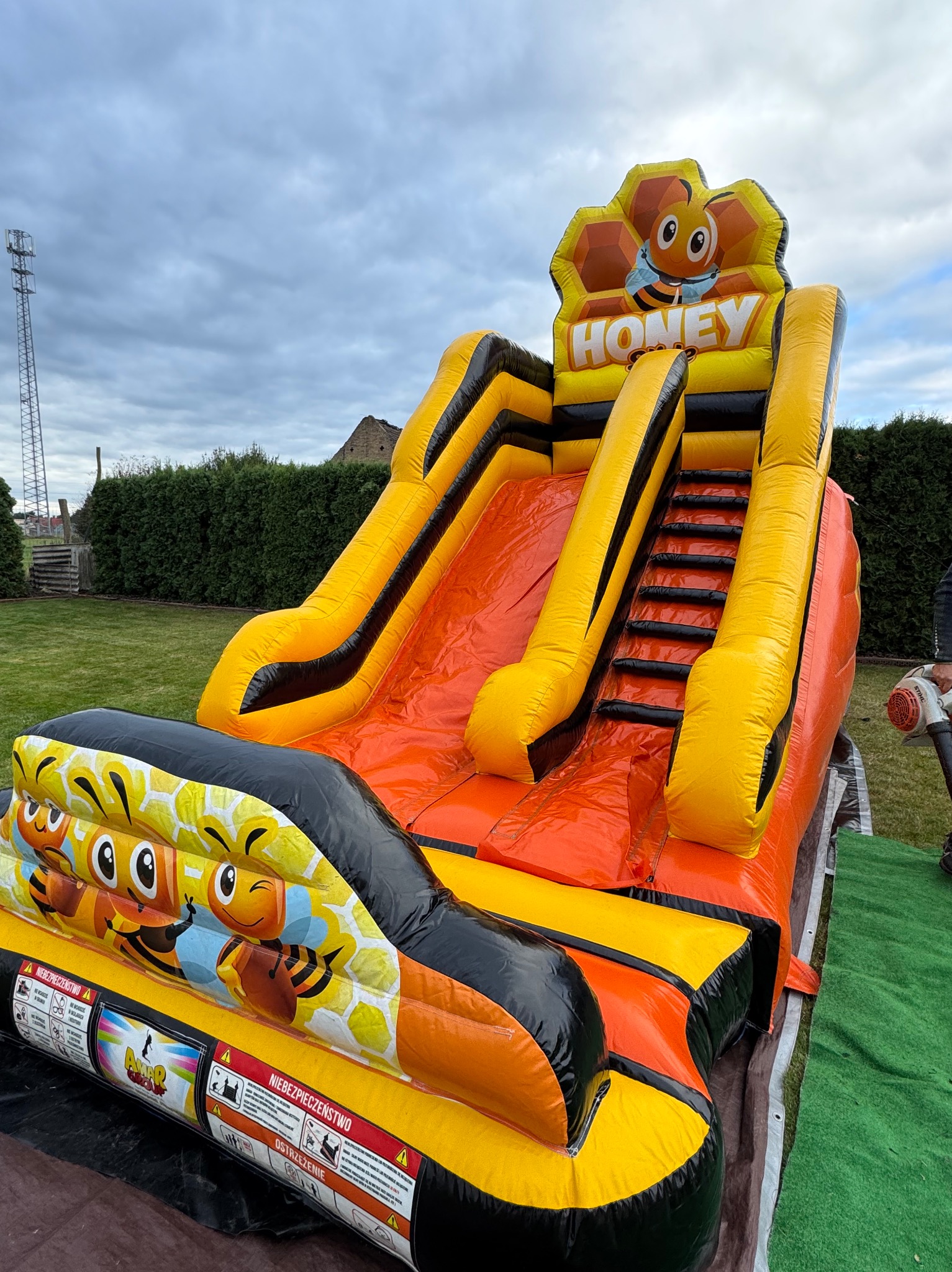 Pomarańczowo-żółta dmuchana zjeżdżalnia z motywem pszczoły na trawniku, widoczna wieża telekomunikacyjna w tle.