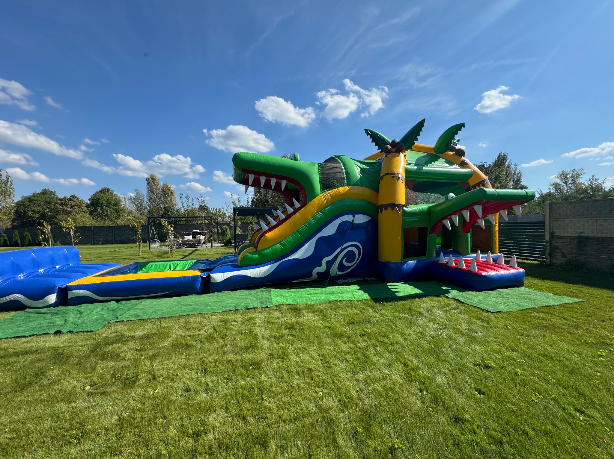 Wielki, nadmuchiwany krokodyl z dwoma zjeżdżalniami i basenem na trawniku, w tle błękitne niebo z chmurami.