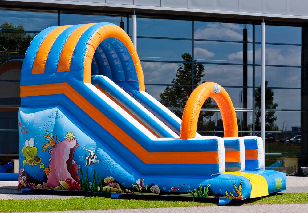 Nadmuchiwania zjeżdżalnia w kolorach niebieskim i pomarańczowym, z motywami podwodnego świata, ustawiona na trawie przed budynkiem z dużymi oknami.