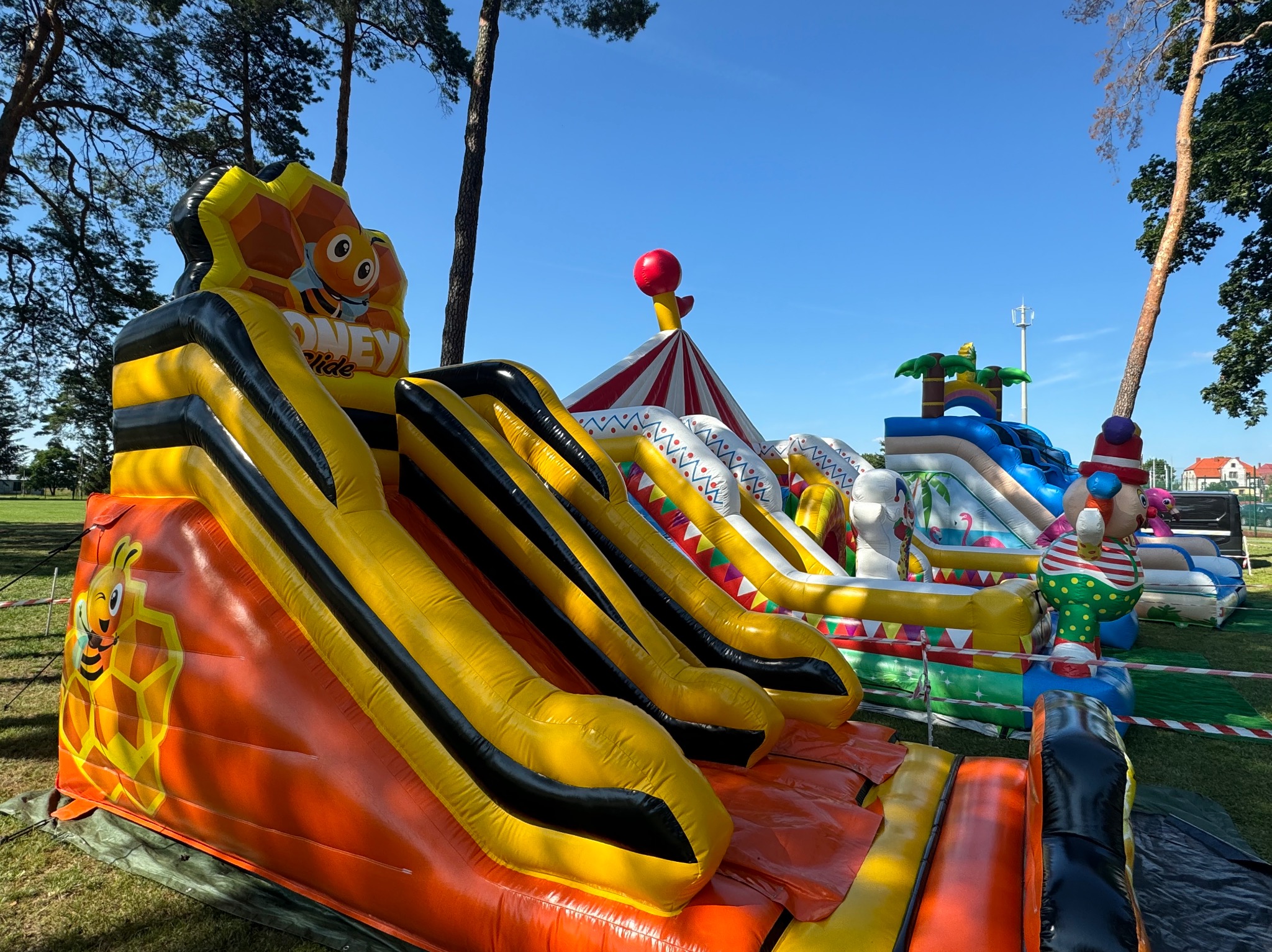 Różnorodne dmuchańce na trawie w słoneczny dzień: zjeżdżalnia z motywem pszczoły, namiot cyrkowy i basen z palmami.