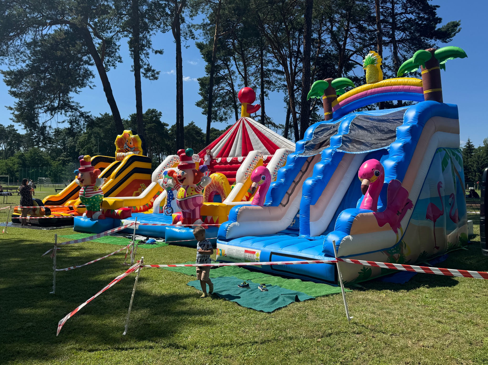 Różnorodne dmuchane zamki i zjeżdżalnie na trawniku, otoczone taśmą ostrzegawczą, z motywami klaunów, dżungli i cyrku, w tle drzewa i niebo.