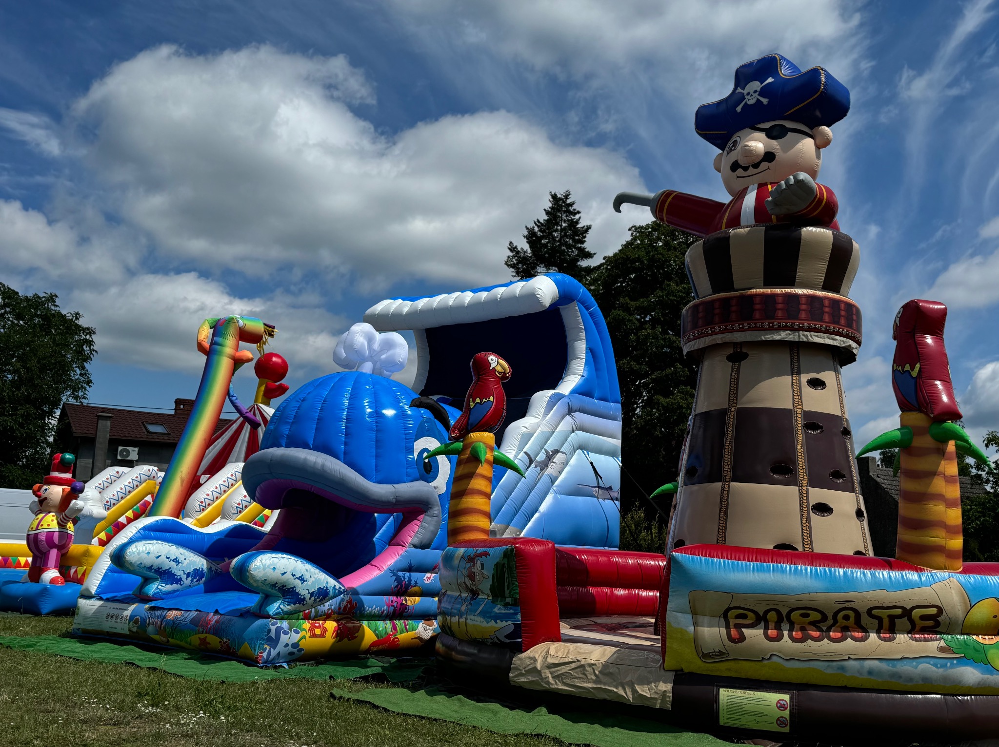 Kompleksowy, kolorowy plac zabaw, w tym dmuchany zamek w kształcie pirata na wieży, zjeżdżalnia w kształcie wieloryba i tęczowa zjeżdżalnia, ustawiony na trawie pod błękitnym niebem z chmurami.