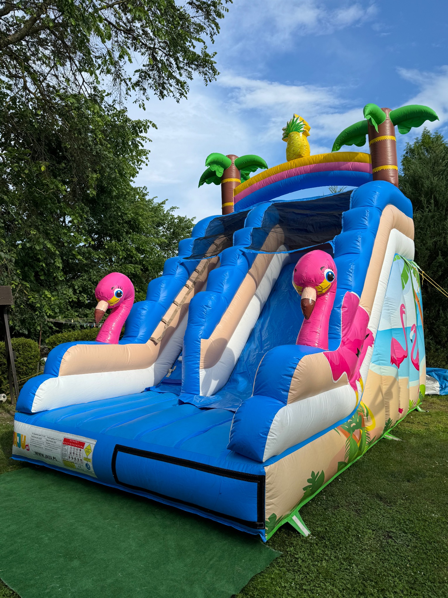 Nadmorski dmuchany plac zabaw ze zjeżdżalnią, ozdobiony palmami, ananasem i różowymi flamingami, ustawiony na trawniku pod błękitnym niebem.