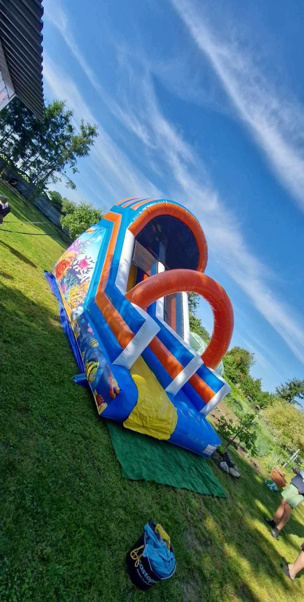 Kolorowa zjeżdżalnia na trawie w Przemysławiu z motywem podwodnego świata, widoczna z boku pod kątem, na tle błękitnego nieba z chmurami.