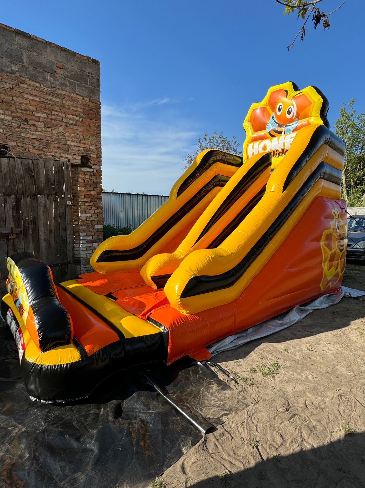 Pomarańczowo-żółta dmuchana zjeżdżalnia w kształcie plastra miodu z wizerunkiem uśmiechniętej pszczoły, ustawiona na zewnątrz obok ceglanego muru i drewnianych wrót.