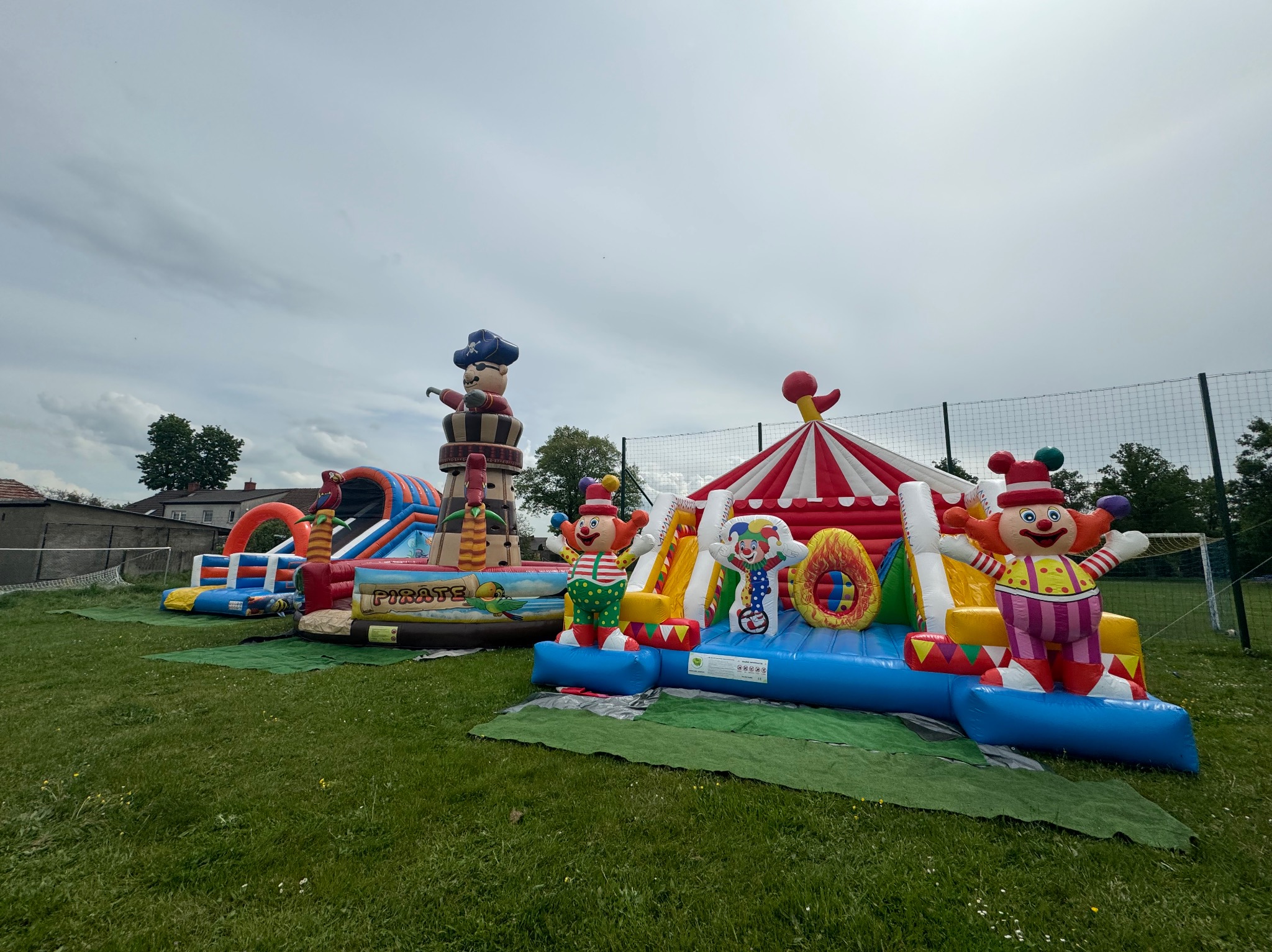 Dwa dmuchane place zabaw na trawie: jeden z motywem pirackim z figurą pirata na szczycie, drugi z motywem cyrkowym z figurami klaunów i namiotem cyrkowym w tle.