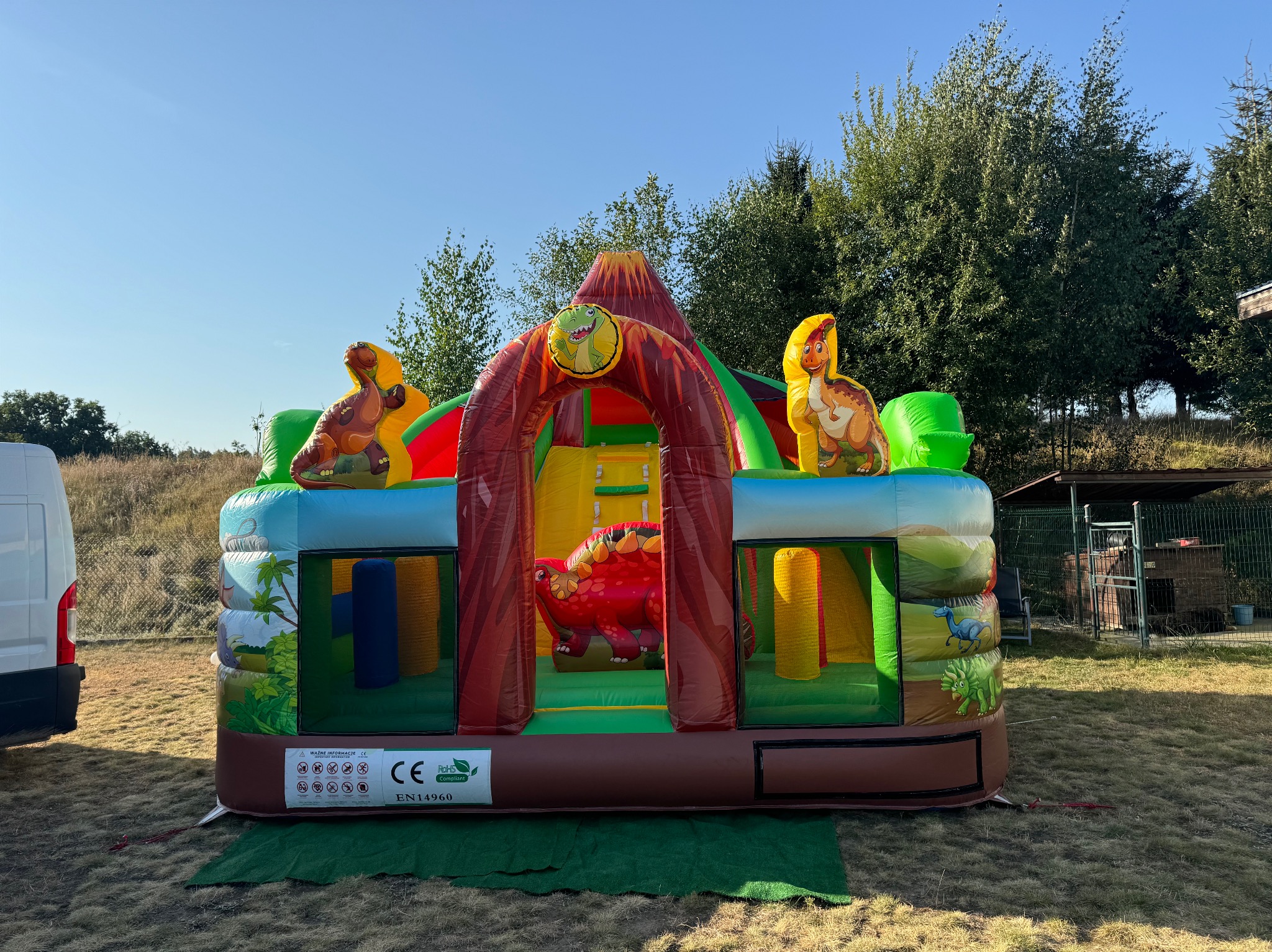 Kolorowy, dmuchany zamek w motywie dinozaurów z wulkanem, ustawiony na trawie w słoneczny dzień, z częściowo widocznym samochodem dostawczym po lewej stronie.