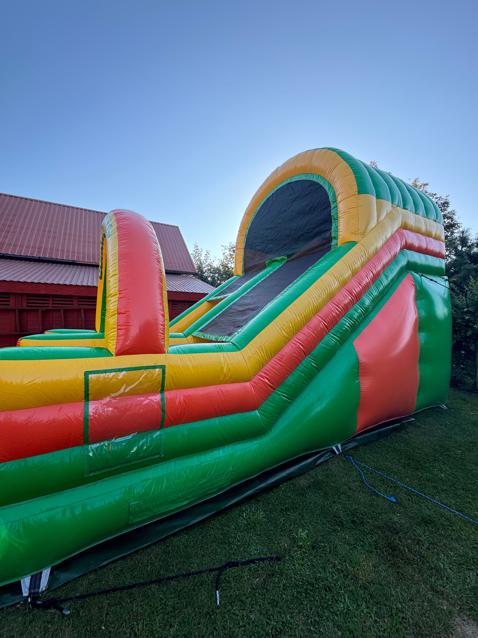 Kolorowa dmuchana zjeżdżalnia z motywem tęczy na trawniku przed czerwonym budynkiem gospodarczym w słoneczny dzień.