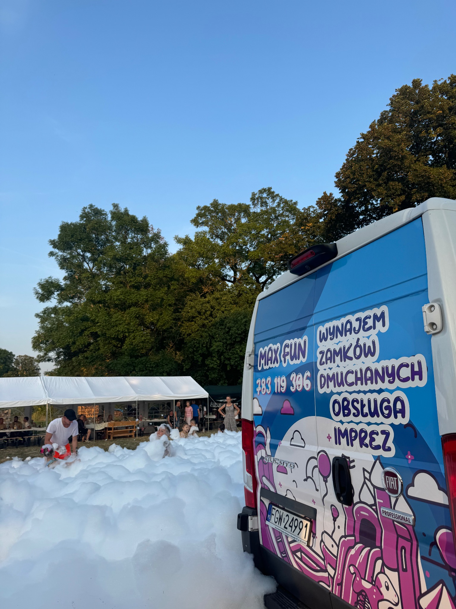 Piana party z animatorem i dziećmi, widoczny samochód z napisem 'Wynajem Zamków Dmuchanych' i numerem telefonu na tle białego namiotu.