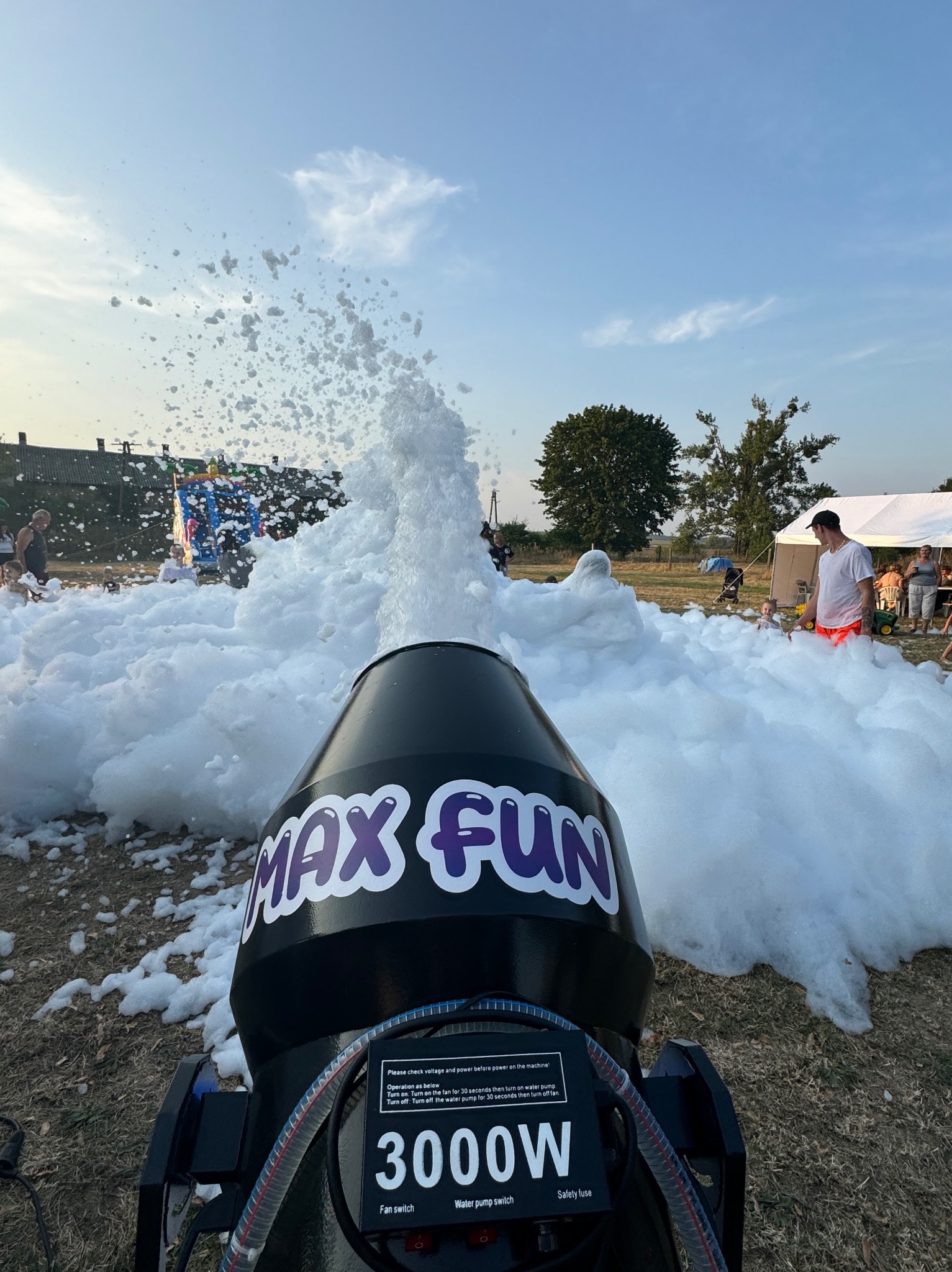 Maszyna do piany z napisem 'MAX FUN' wyrzucająca strumień piany na imprezie plenerowej, w tle bawiący się ludzie i dmuchany zamek.