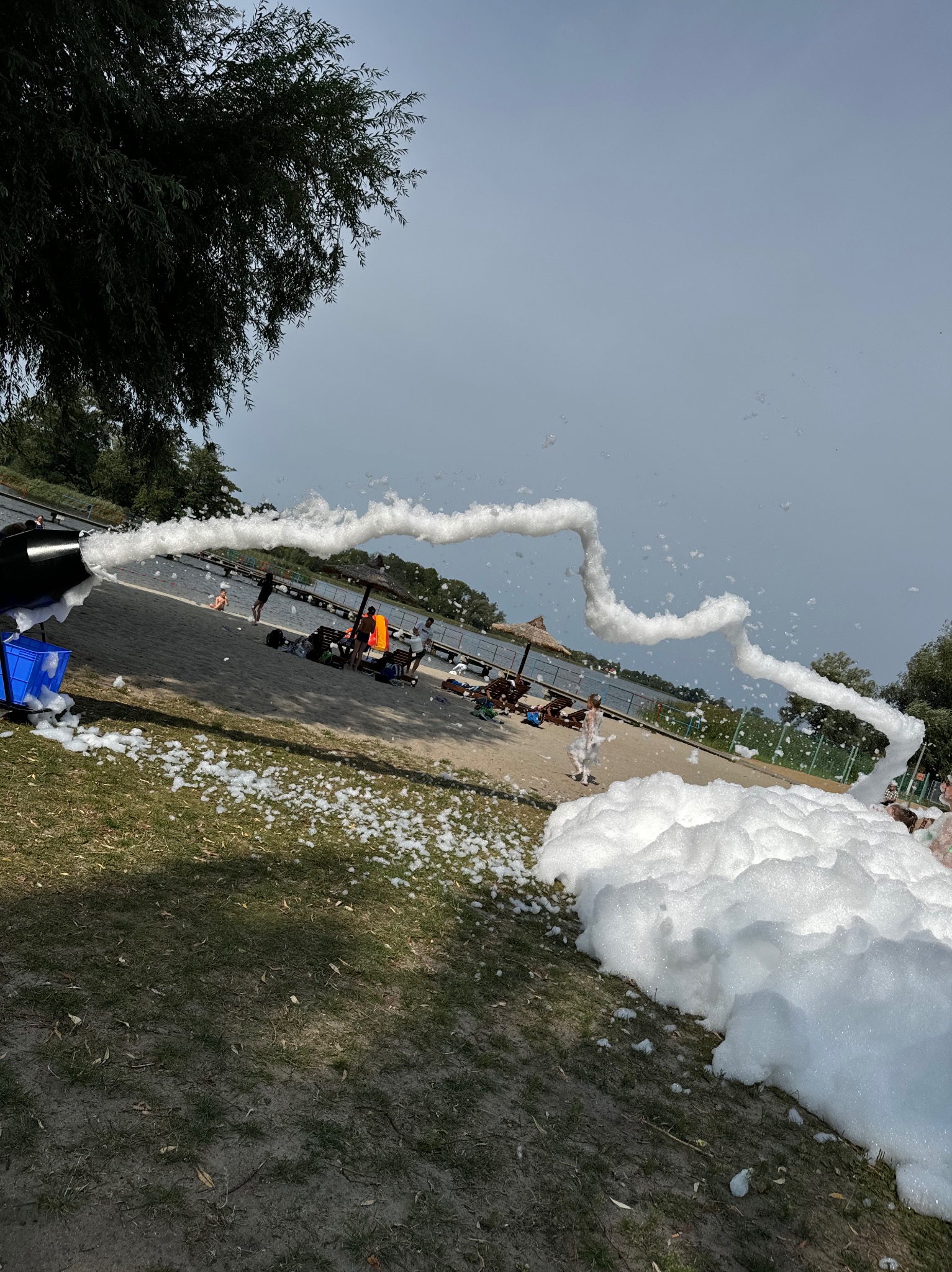 Piana imprezowa wydobywająca się z czarnej maszyny na tle piaszczystej plaży z ludźmi i zbiornika wodnego.