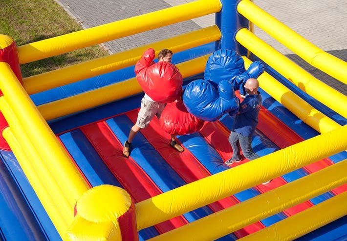 Dwóch mężczyzn wewnątrz dmuchanego ringu bokserskiego z ogromnymi, kolorowymi rękawicami, w trakcie zabawy.