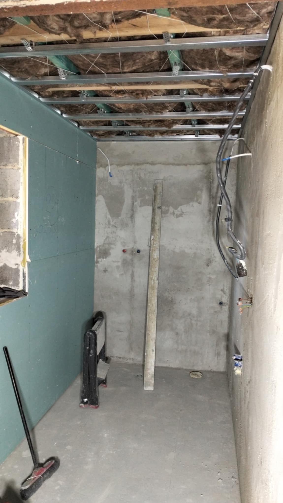 Wnętrze w trakcie prac: zamontowany stelaż sufitu podwieszanego z izolacją, częściowo obłożona ściana płytą gipsowo-kartonową, widoczne instalacje elektryczne i pionowa poziomica oparta o surową...