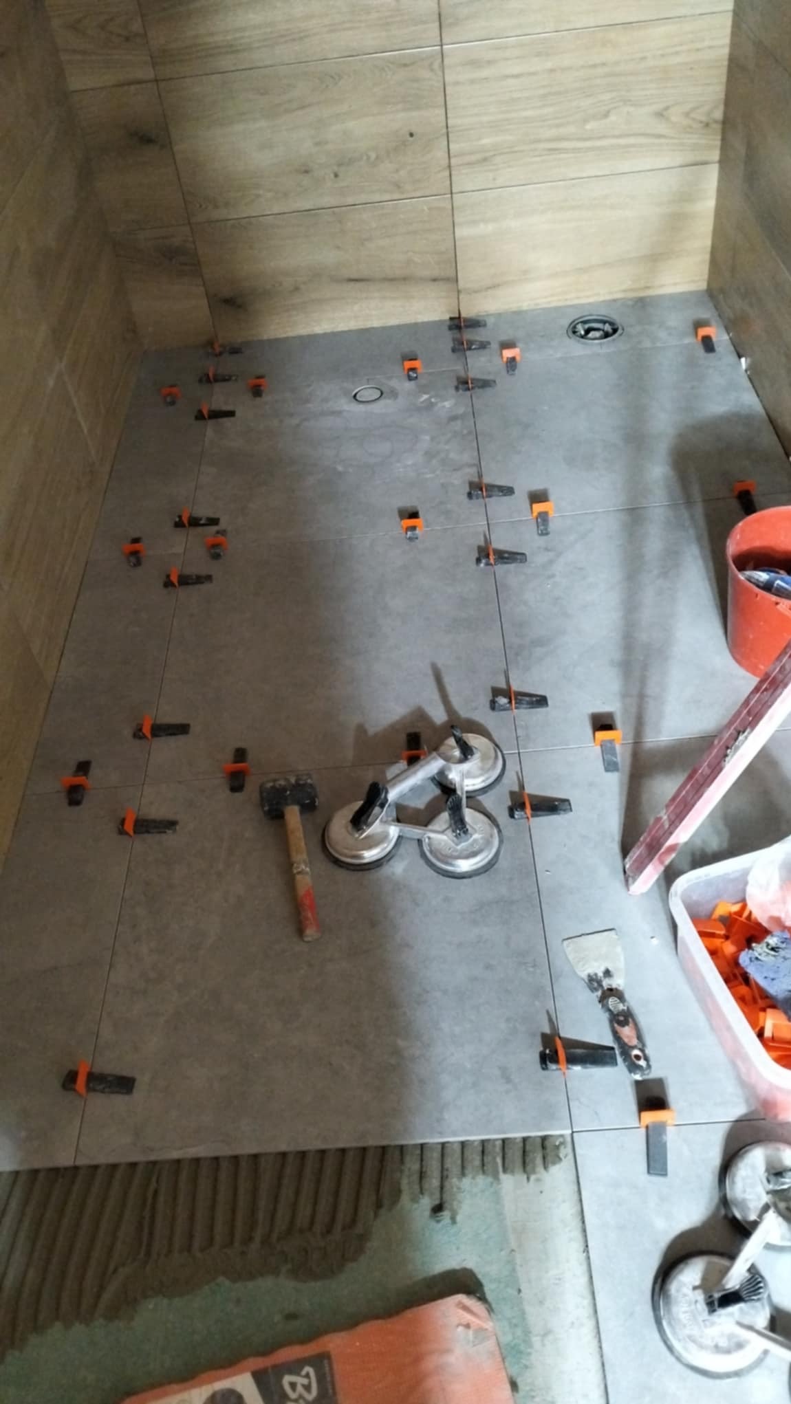 Układanie szarych płytek podłogowych w łazience z drewnopodobnymi płytkami ściennymi, widoczne przyssawki do płytek, młotek, szpachelka i system poziomowania płytek z pomarańczowymi klipsami.