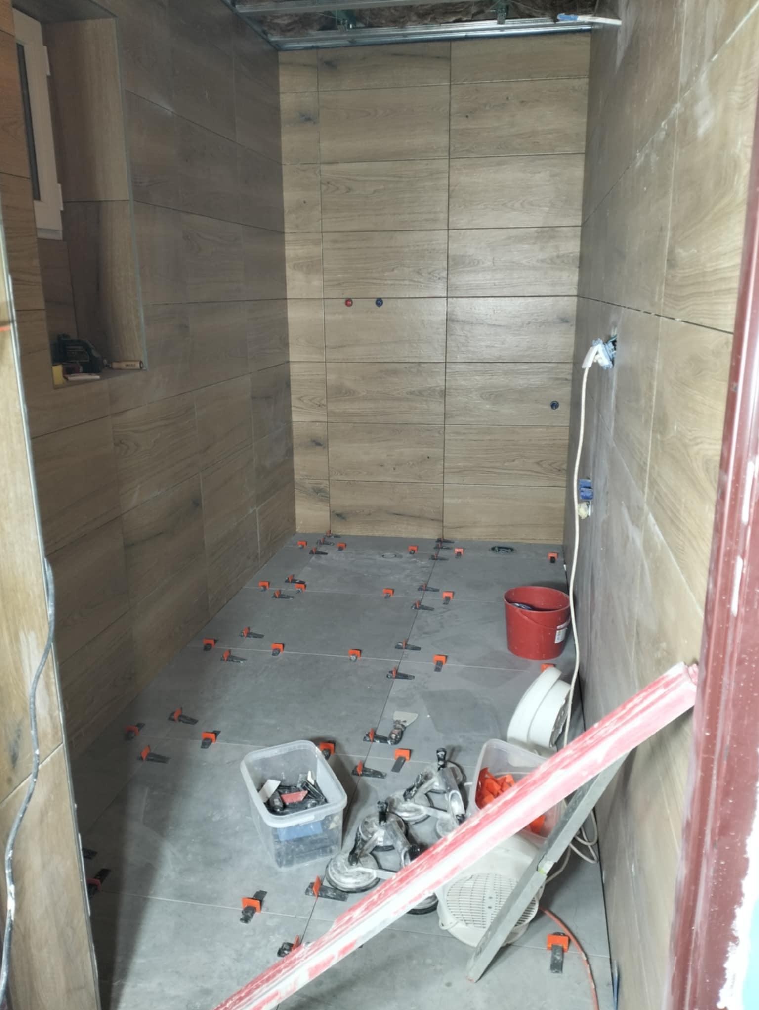 Wnętrze łazienki w trakcie prac, widoczne płytki imitujące drewno na ścianach, świeżo ułożone szare płytki na podłodze z systemem poziomowania, wiadro, poziomica i narzędzia glazurnicze.