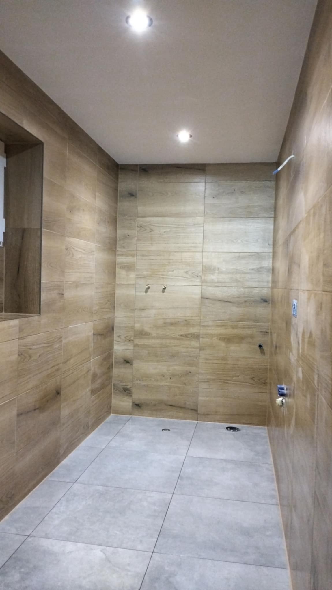 Ukończona łazienka z drewnopodobnymi płytkami ściennymi i szarą podłogą, widoczne dwa punkty świetlne w suficie i przygotowane punkty podłączeń hydraulicznych.