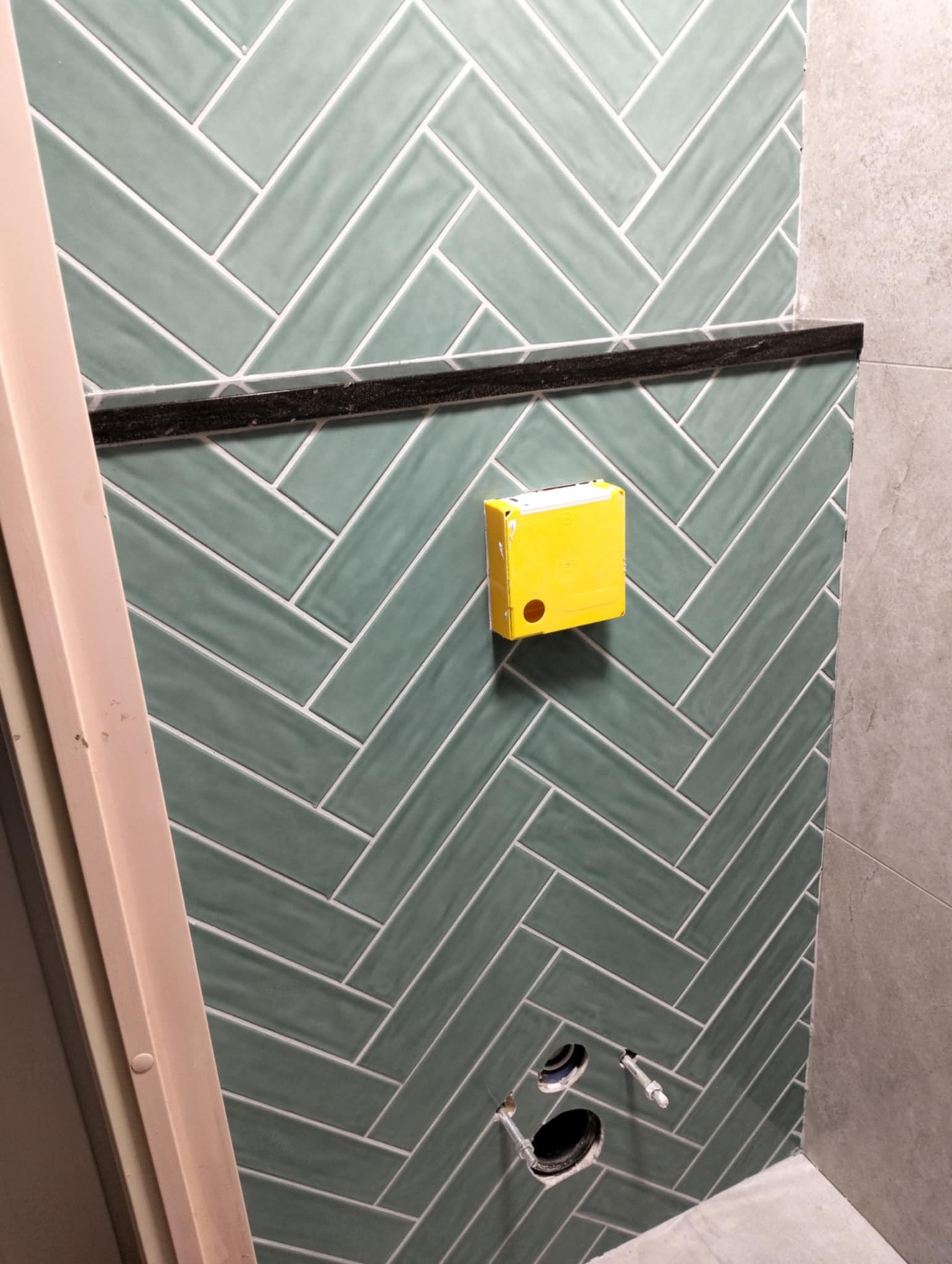 Ściana łazienki wyłożona zielonymi, podłużnymi płytkami ułożonymi w jodełkę, z czarnym pasem dekoracyjnym i żółtą puszką elektryczną, widoczne otwory na instalację wodną.