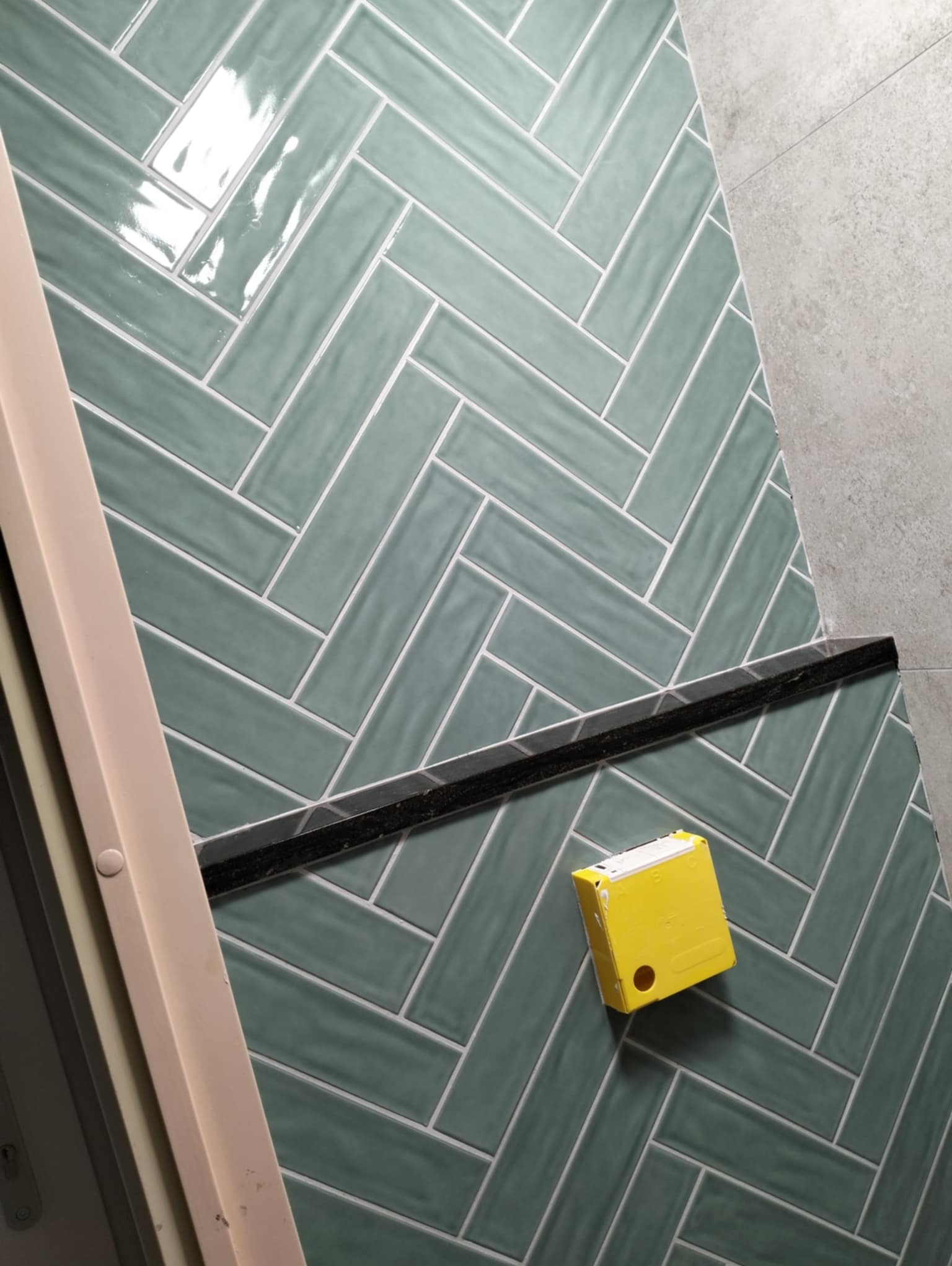 Ściana łazienki wyłożona błyszczącymi, turkusowymi płytkami ułożonymi w jodełkę, z widoczną listwą dekoracyjną i żółtą miarką zwijaną.