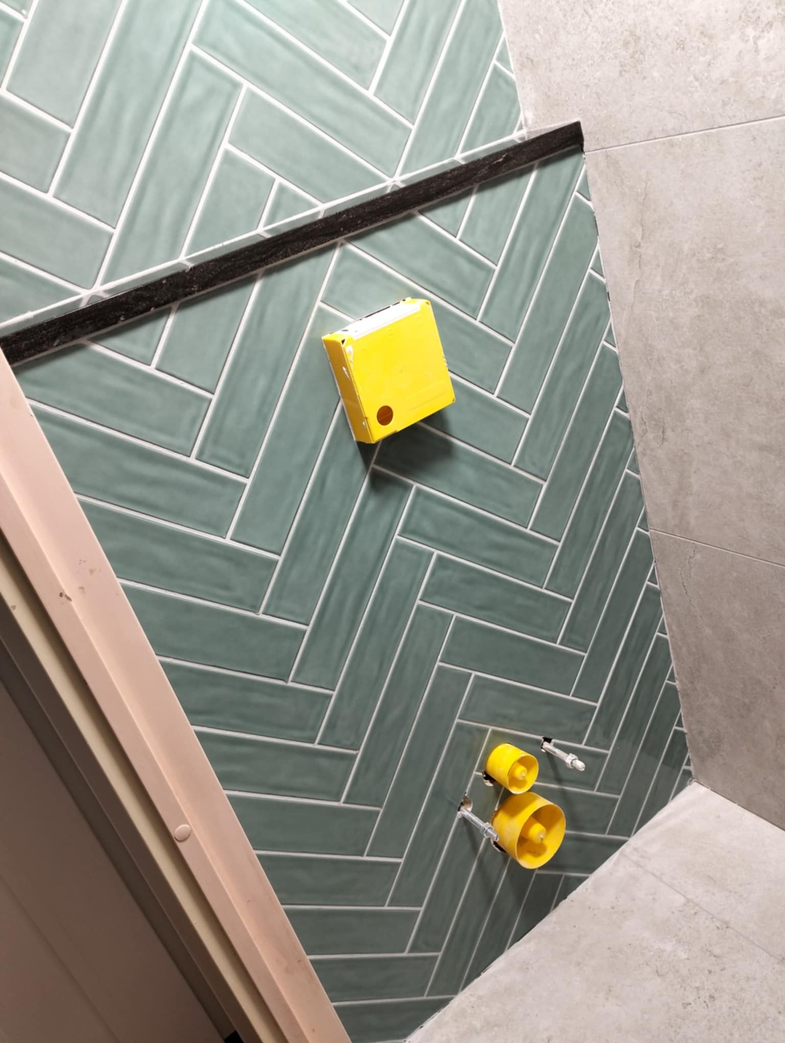 Ściana w Piątkowej wyłożona zielonymi płytkami ułożonymi w jodełkę, z widocznymi elementami instalacji wodnej i czarną listwą dekoracyjną.