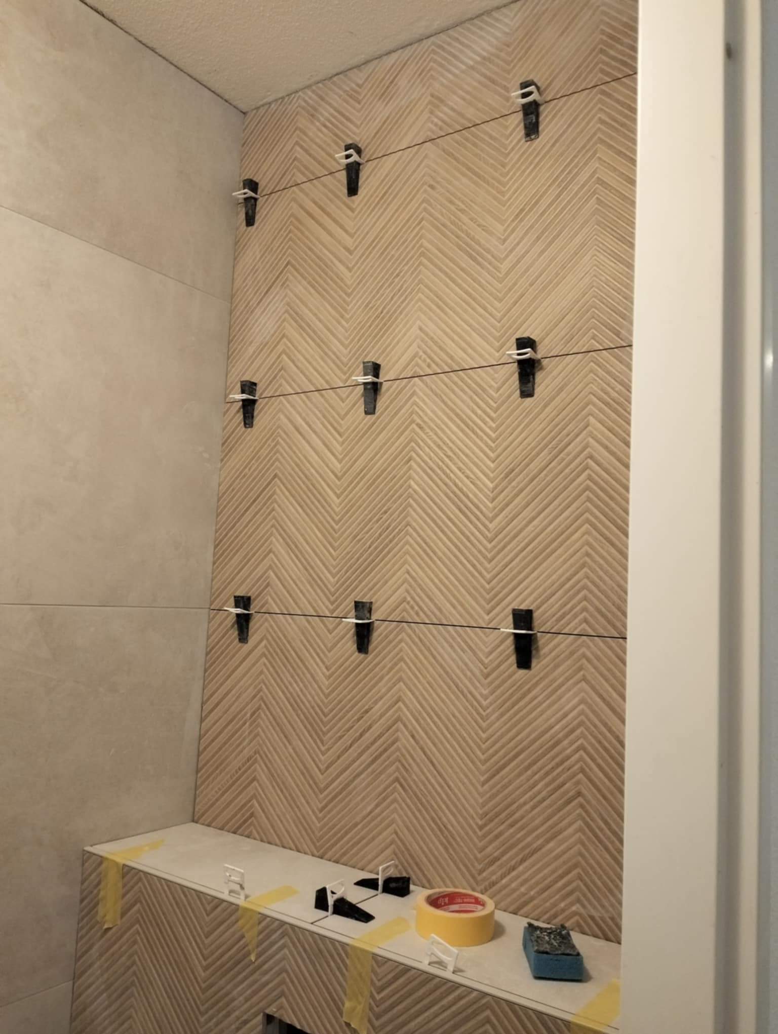 Ściana łazienki w trakcie układania płytek w jodełkę, z użyciem klipsów poziomujących i sznurka, widoczna półka z taśmą malarską i gąbką.