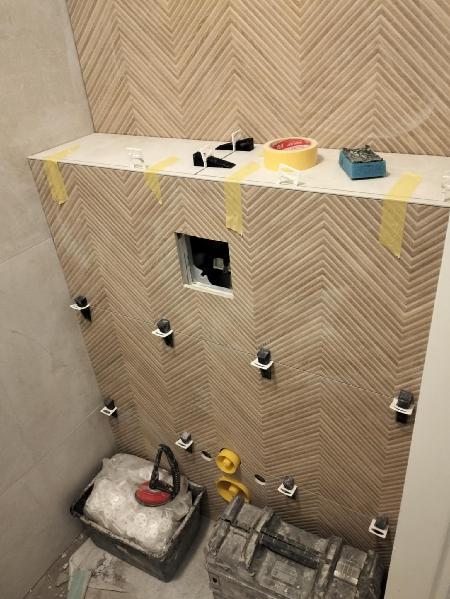 Montaż płytek w łazience z wzorem jodełki, widoczne narzędzia glazurnicze, system poziomowania płytek i otwór na instalację sanitarną.