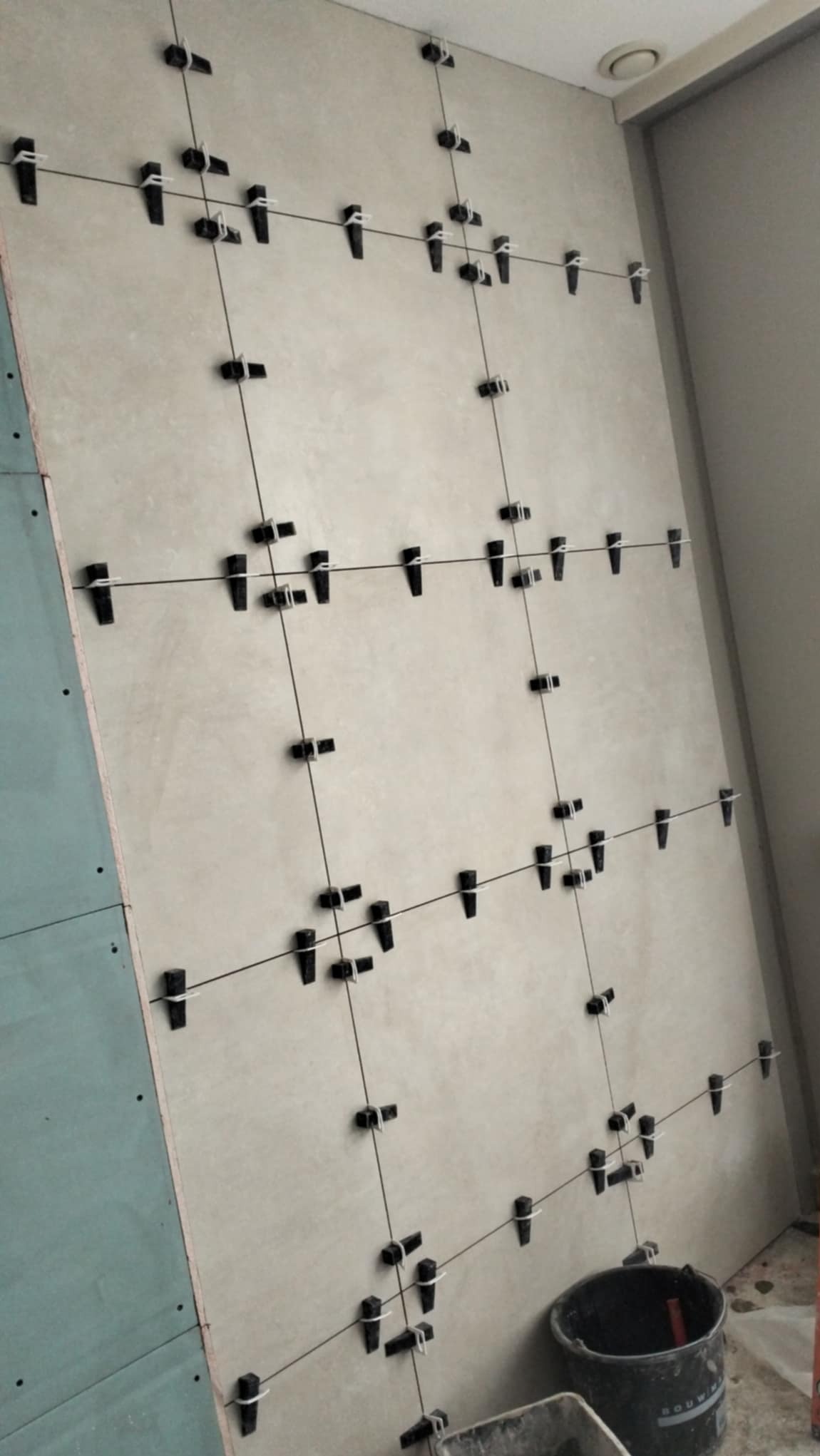 Ściana w trakcie układania płytek z użyciem systemu poziomowania: klipsy i kliny rozmieszczone w regularnych odstępach, widoczna płyta kartonowo-gipsowa po lewej stronie oraz wiadro z zaprawą...