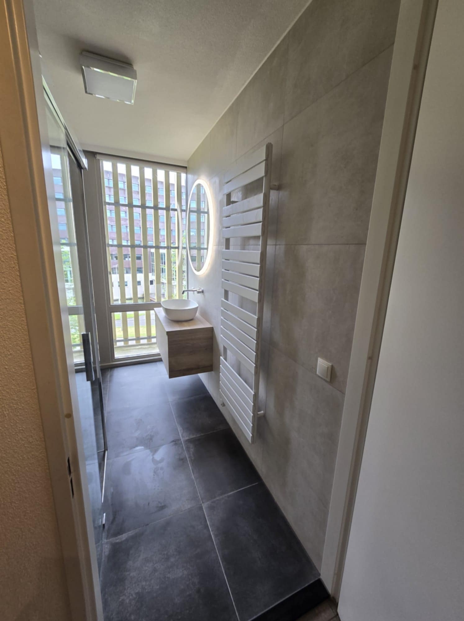 Nowoczesna łazienka z szarymi płytkami, podświetlanym okrągłym lustrem, drewnianą szafką pod umywalką, białym grzejnikiem drabinkowym i ciemnymi płytkami podłogowymi, widok zza otwartych drzwi.