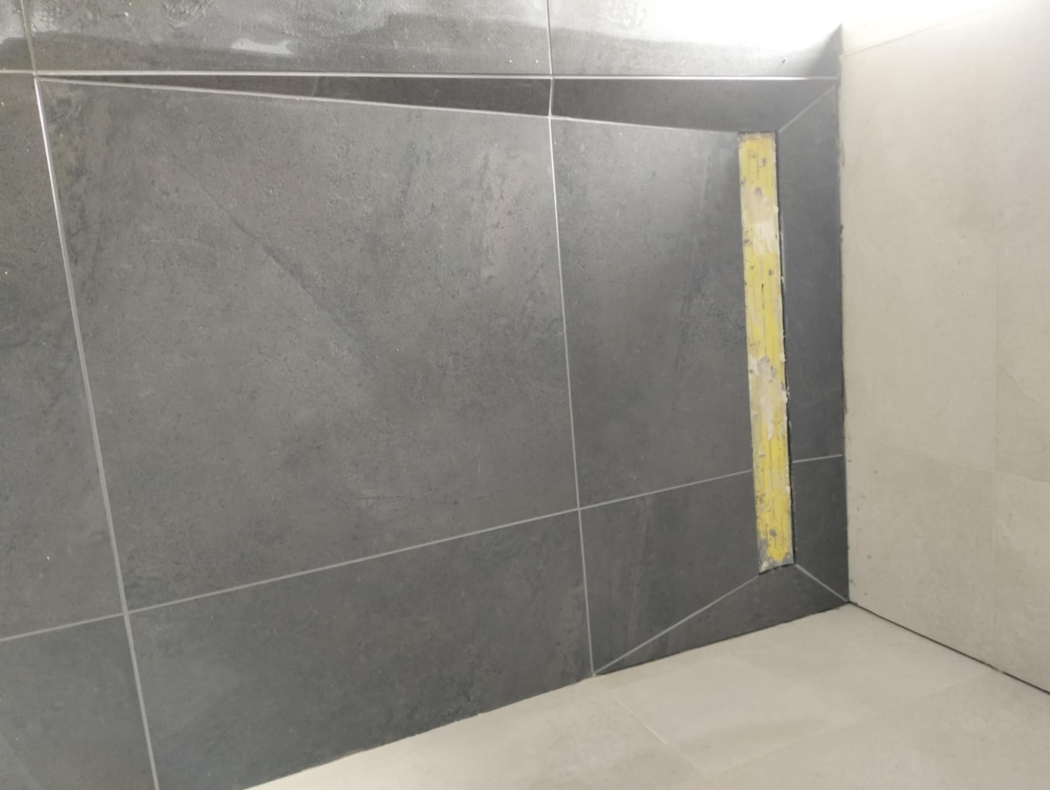 Fragment ściany łazienki wyłożonej ciemnymi, matowymi płytkami z fugami w kolorze szarym, z widocznym narożnikiem wykończonym żółtą taśmą malarską zabezpieczającą.