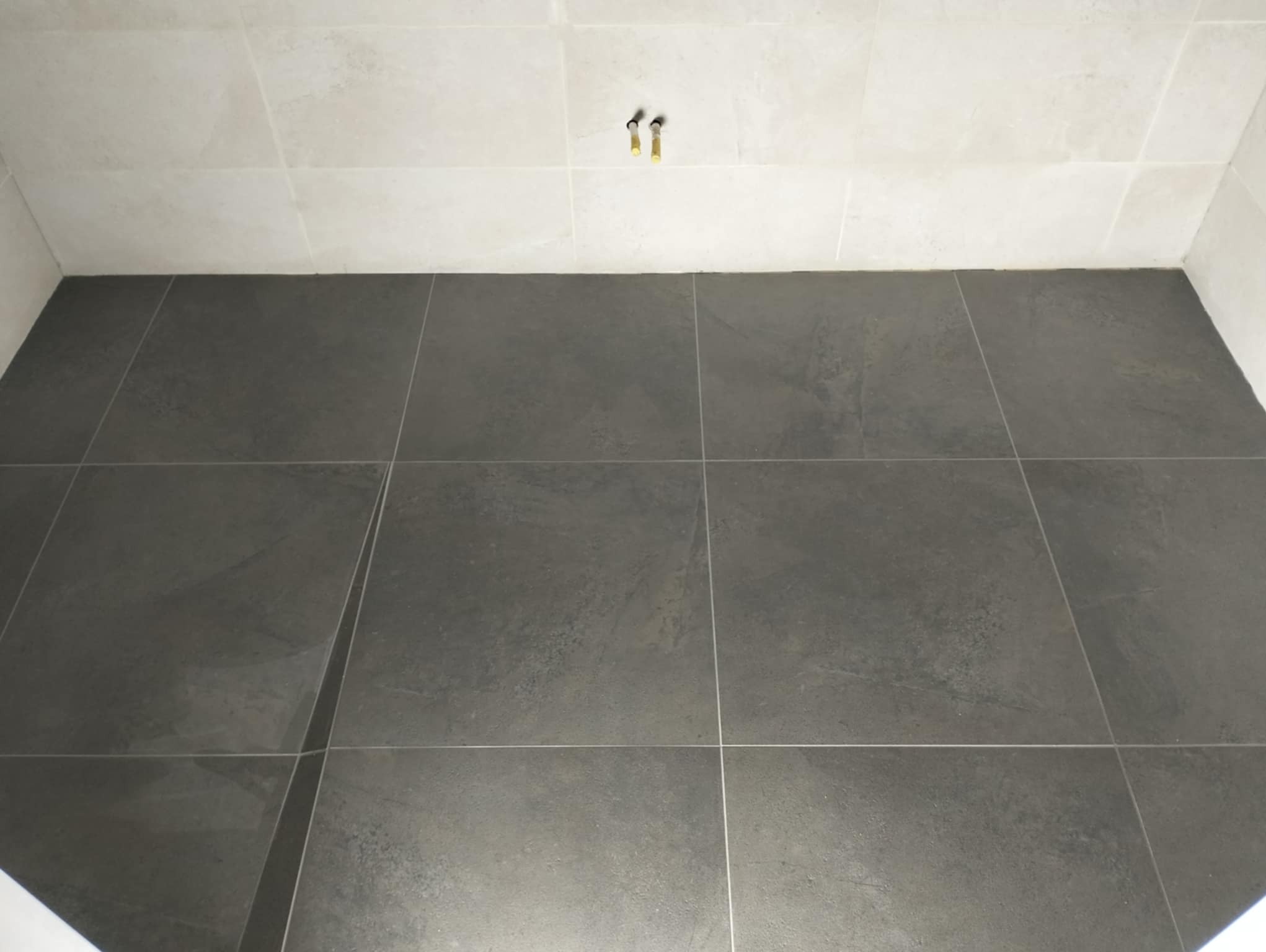 Ułożone ciemnoszare płytki podłogowe w łazience, jasne płytki ścienne, wystające rury do armatury.