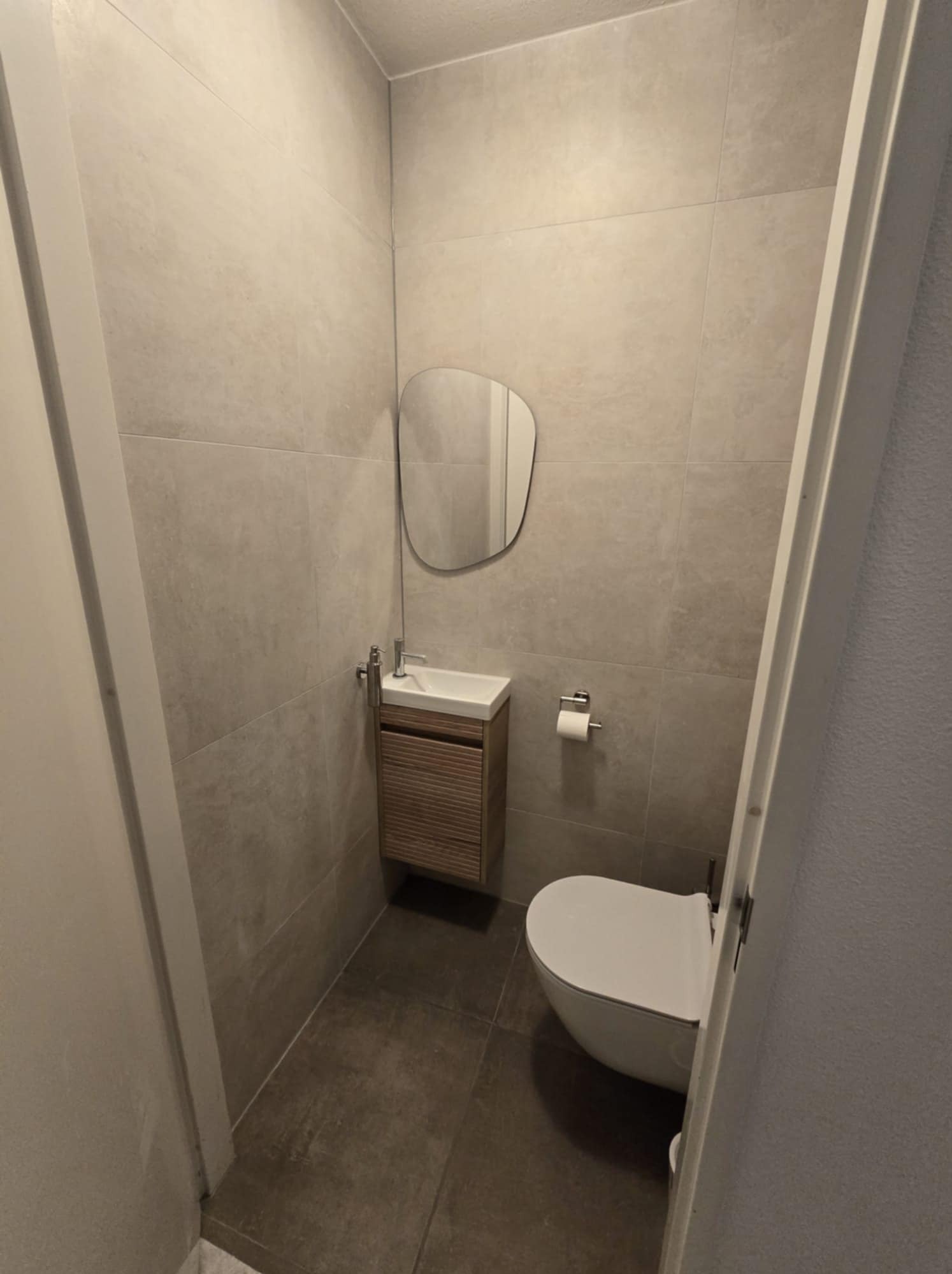 Nowoczesna, minimalistyczna łazienka z beżowymi płytkami, wiszącą toaletą i umywalką z szafką z lamelami, oświetlona neutralnym światłem.