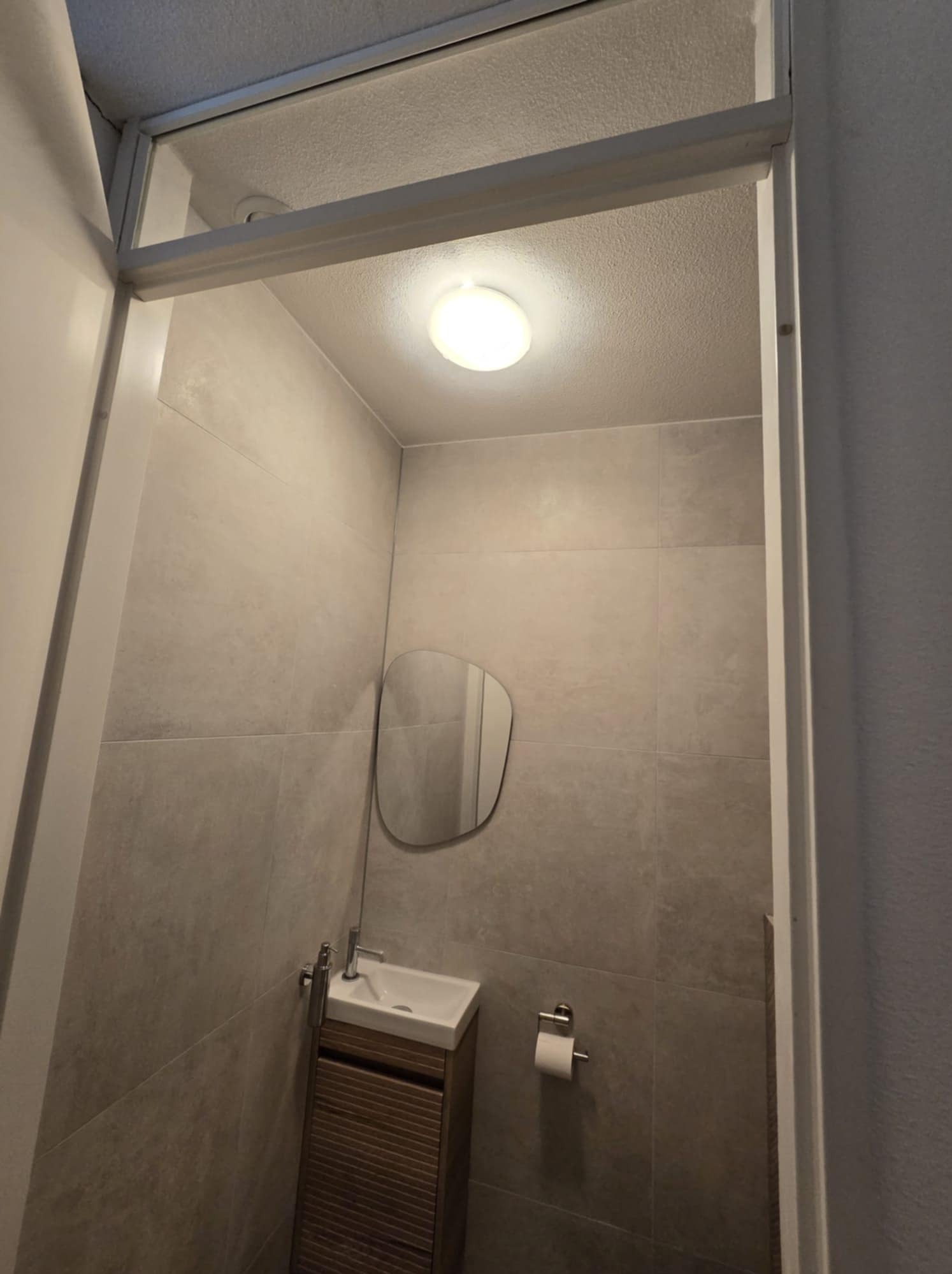 Nowoczesna, minimalistyczna łazienka wykończona w szarych płytkach, z podświetlanym sufitem, małą umywalką z szafką z lamelami oraz asymetrycznym lustrem.