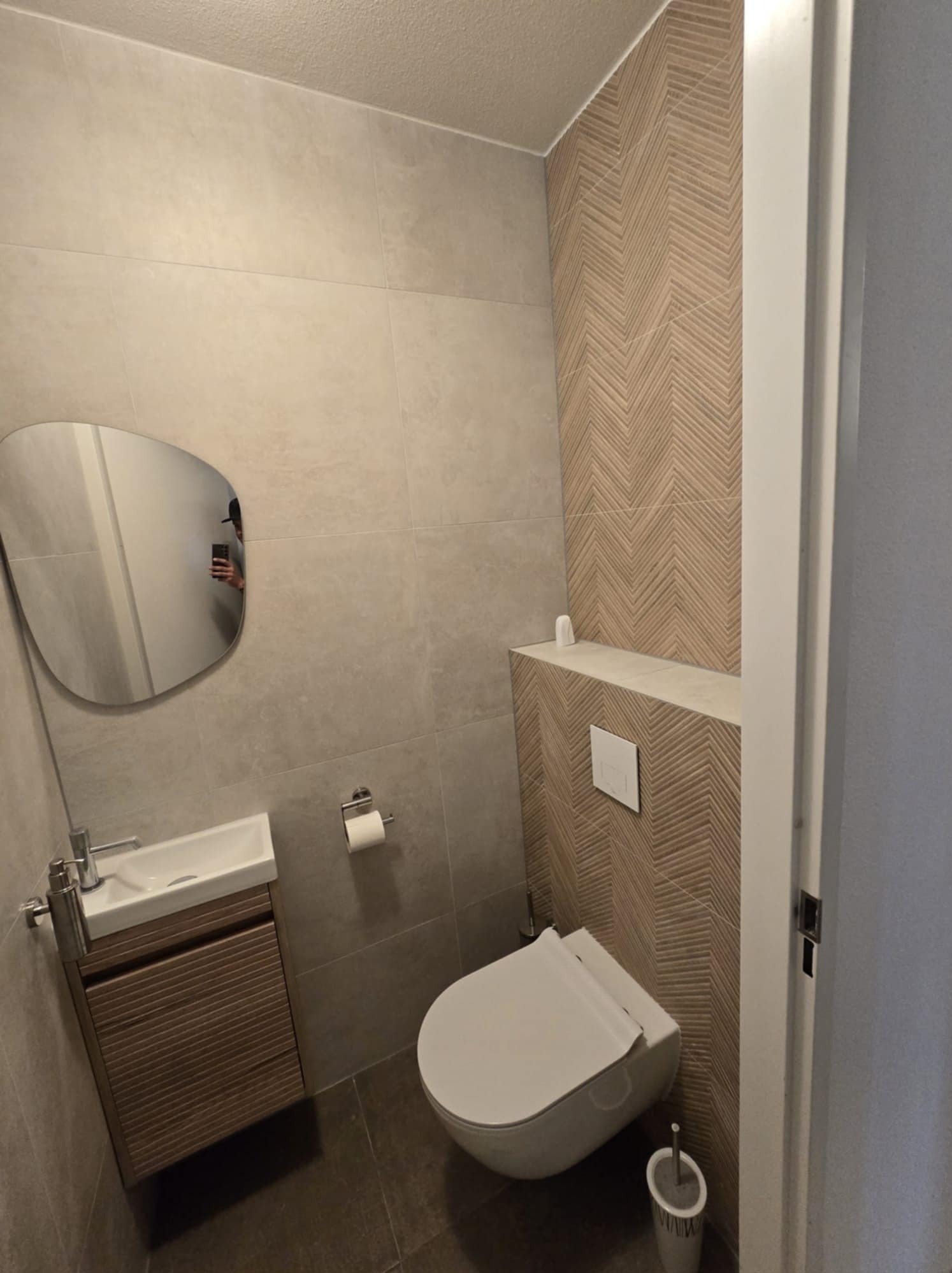 Nowoczesna łazienka z beżowymi płytkami imitującymi drewno ułożonymi w jodełkę na jednej ścianie, wisząca toaleta z wbudowanym przyciskiem spłukującym, asymetryczne lustro odbijające osobę robiącą...