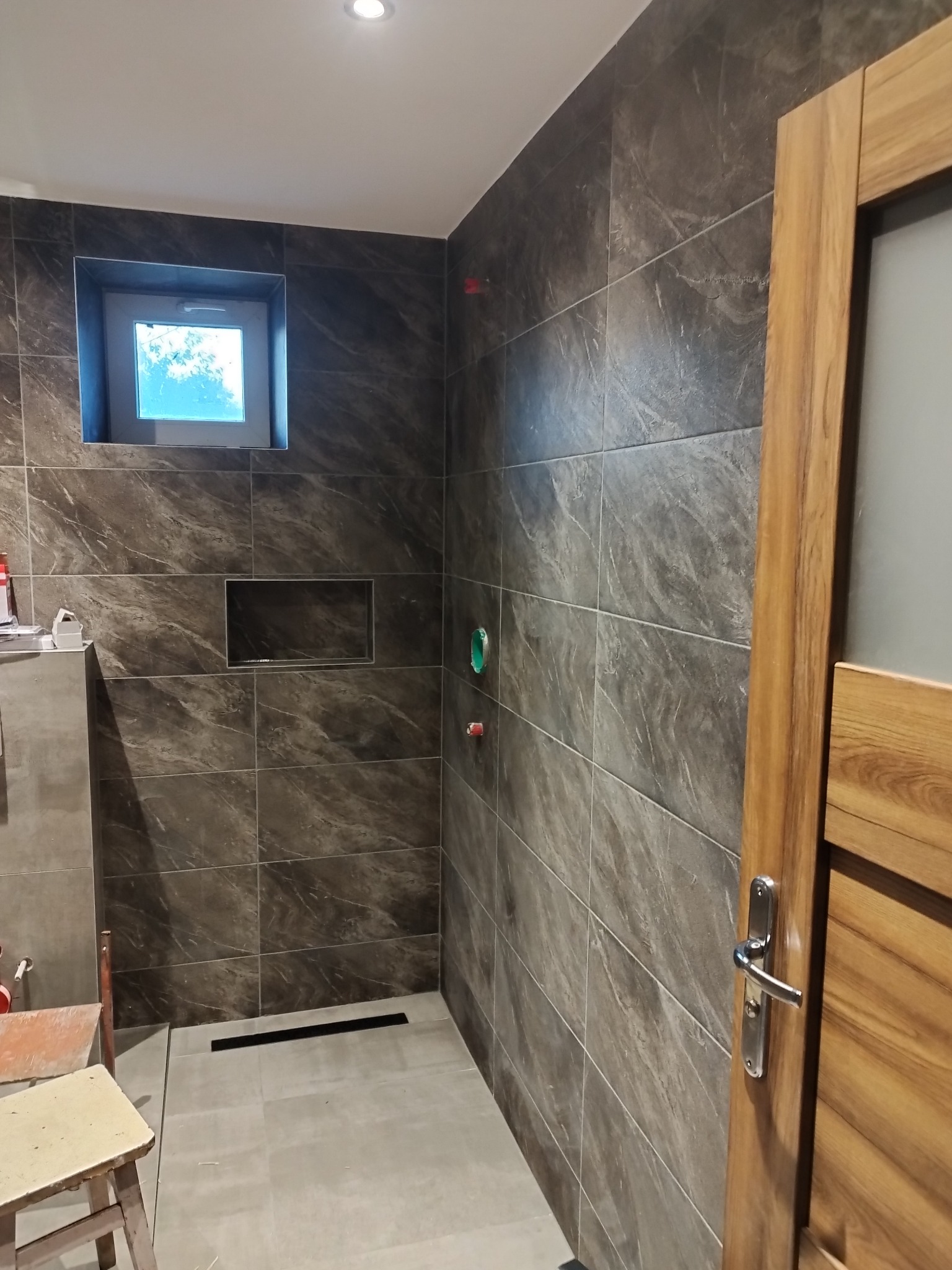 Nowoczesna łazienka z szarymi płytkami imitującymi marmur, wąskim oknem w ramie i drewnianymi drzwiami z chromowaną klamką, widoczny odpływ liniowy w podłodze i półka w ścianie.