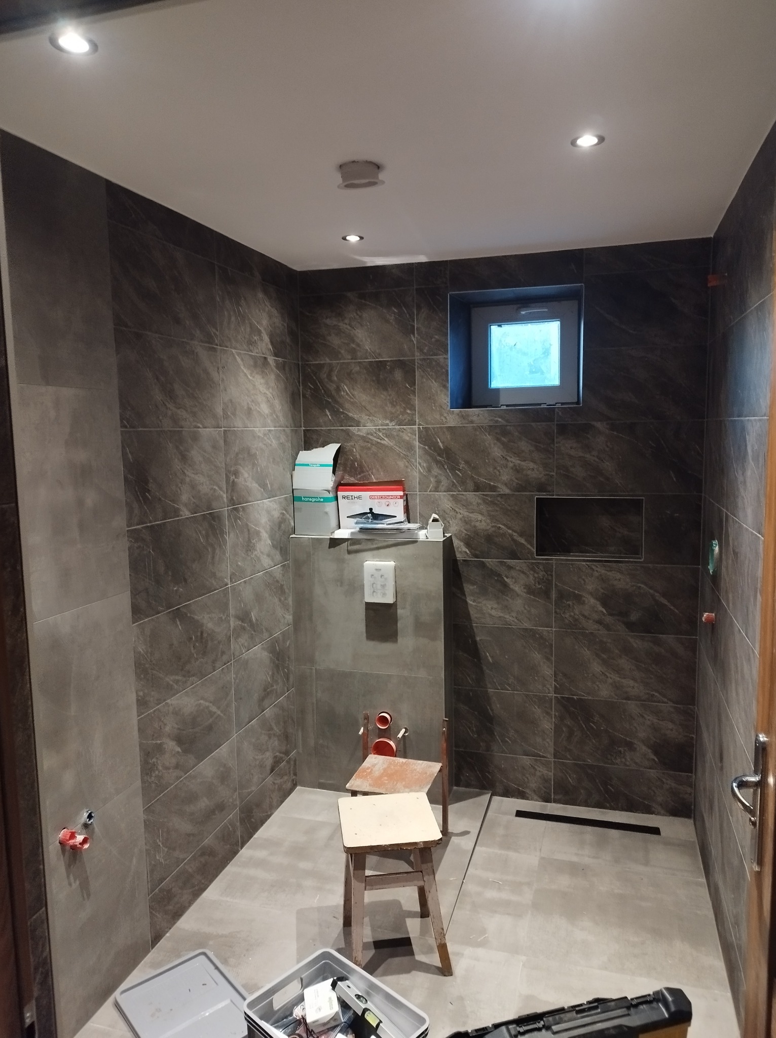 Wnętrze łazienki w trakcie prac wykończeniowych, widoczne płytki imitujące marmur na ścianach i podłodze, fragment ściany z oknem, zamontowany odpływ liniowy, skrzynka z narzędziami na podłodze...