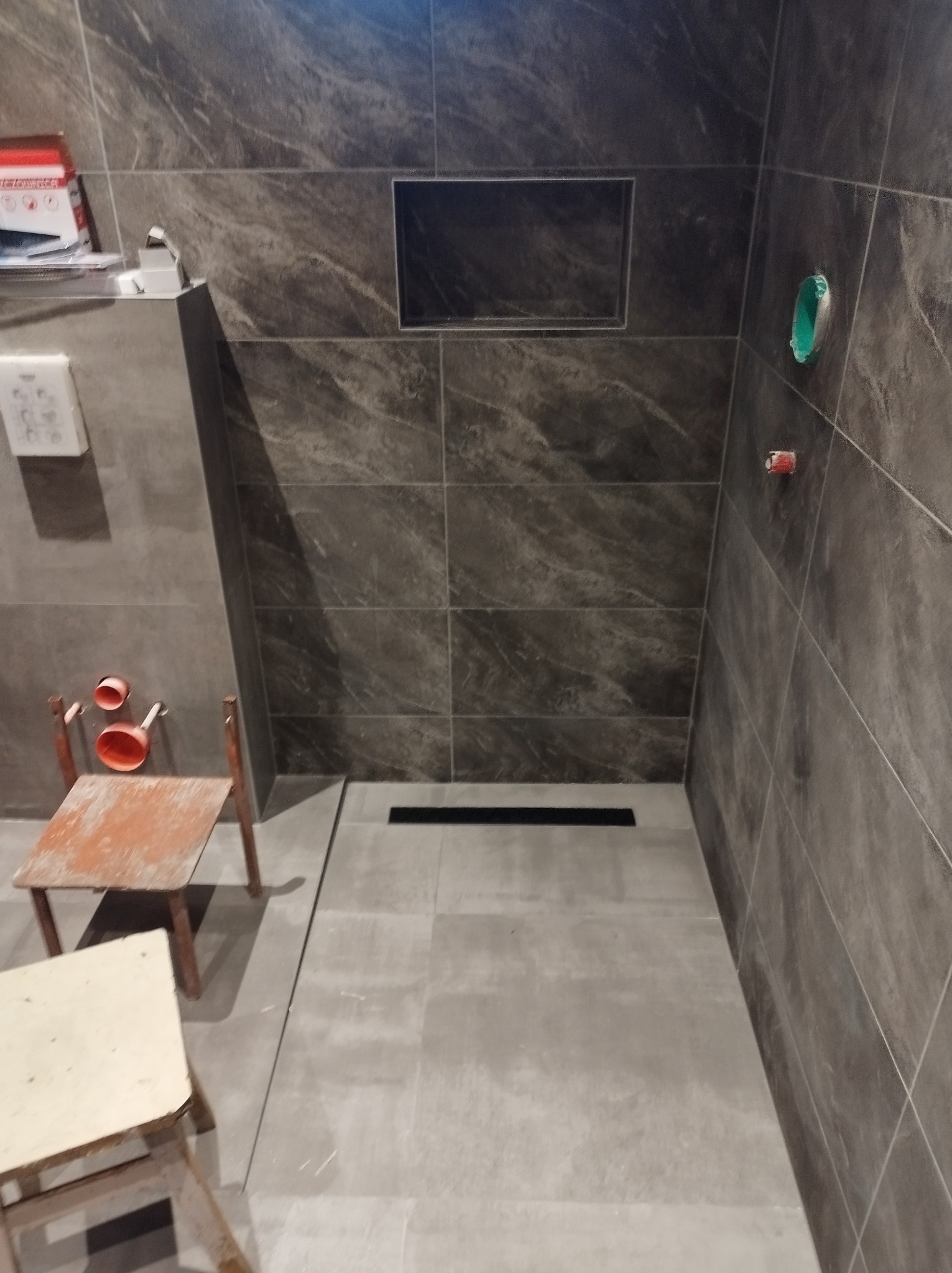 Fragment łazienki w trakcie wykańczania: ciemne, marmurkowe płytki na ścianach, zamontowany odpływ liniowy w podłodze, widoczne przyłącza wodne, prowizoryczne krzesła robocze.