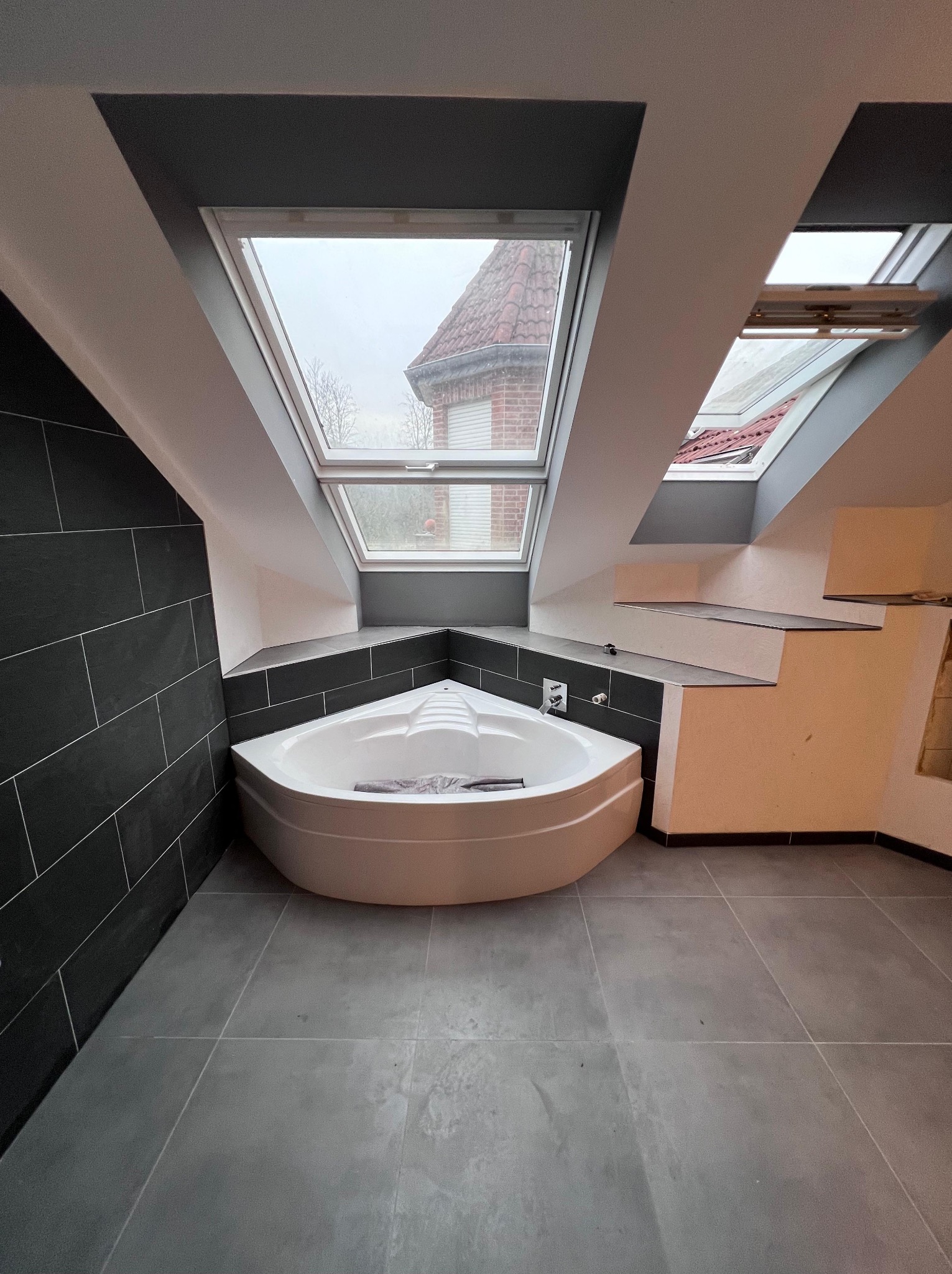 Wykończona łazienka na poddaszu ze skośnym dachem i oknami dachowymi, narożna wanna z hydromasażem zabezpieczona folią, ściany wyłożone ciemnymi płytkami imitującymi kamień, szare płytki podłogowe.