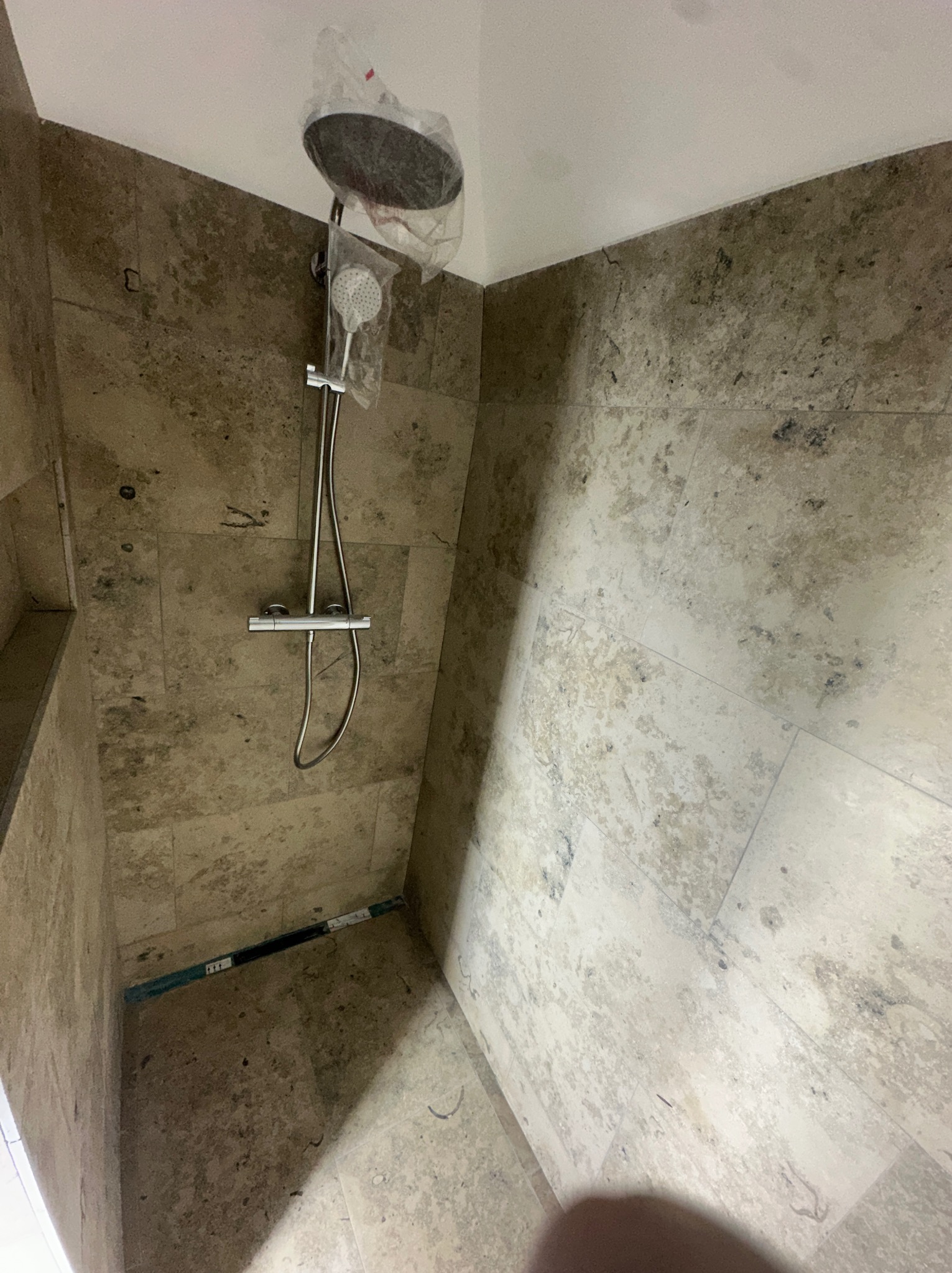 Niedokończona kabina prysznicowa z beżowymi płytkami imitującymi kamień, nowa bateria prysznicowa z deszczownicą w folii ochronnej, widoczny odpływ liniowy w podłodze.
