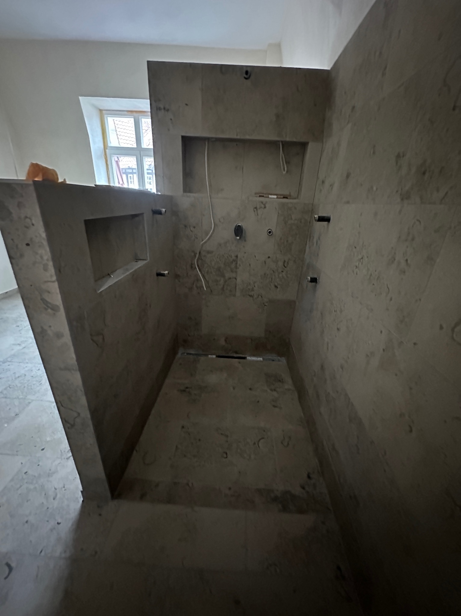 Surowa łazienka w trakcie wykończenia, widoczne płytki w odcieniach szarości i beżu, przygotowane pod instalację armatury, z wnękami w ścianach i oknem w tle.