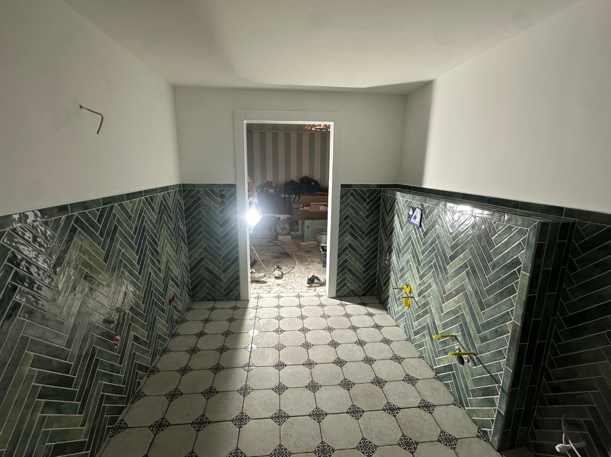 Wnętrze łazienki w trakcie prac wykończeniowych, z zieloną glazurą ułożoną w jodełkę na ścianach, wzorzystą podłogą i widocznymi elementami instalacji. W tle otwarte drzwi do pomieszczenia...