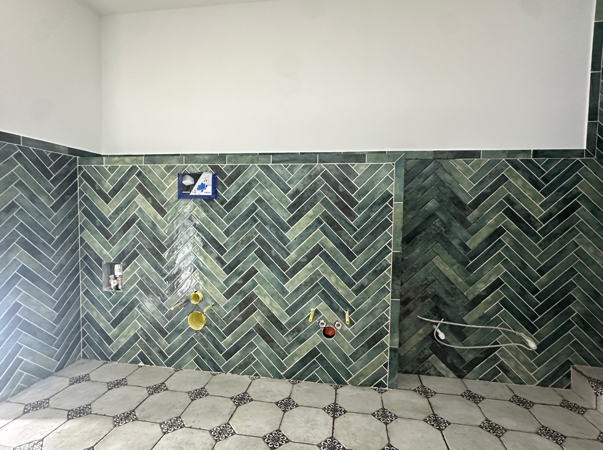 Wnętrze łazienki w trakcie prac wykończeniowych: ściany wyłożone zieloną, błyszczącą glazurą ułożoną w jodełkę, podłoga z płytek w stylu retro, widoczne instalacje wodno-kanalizacyjne.