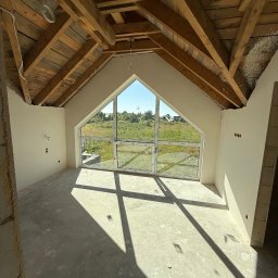Tynkib - Wnętrze poddasza z drewnianą konstrukcją dachu i oknem szczytowym. Ściany po tynkowaniu, widoczne instalacje elektryczne. Na podłodze cień od okna.