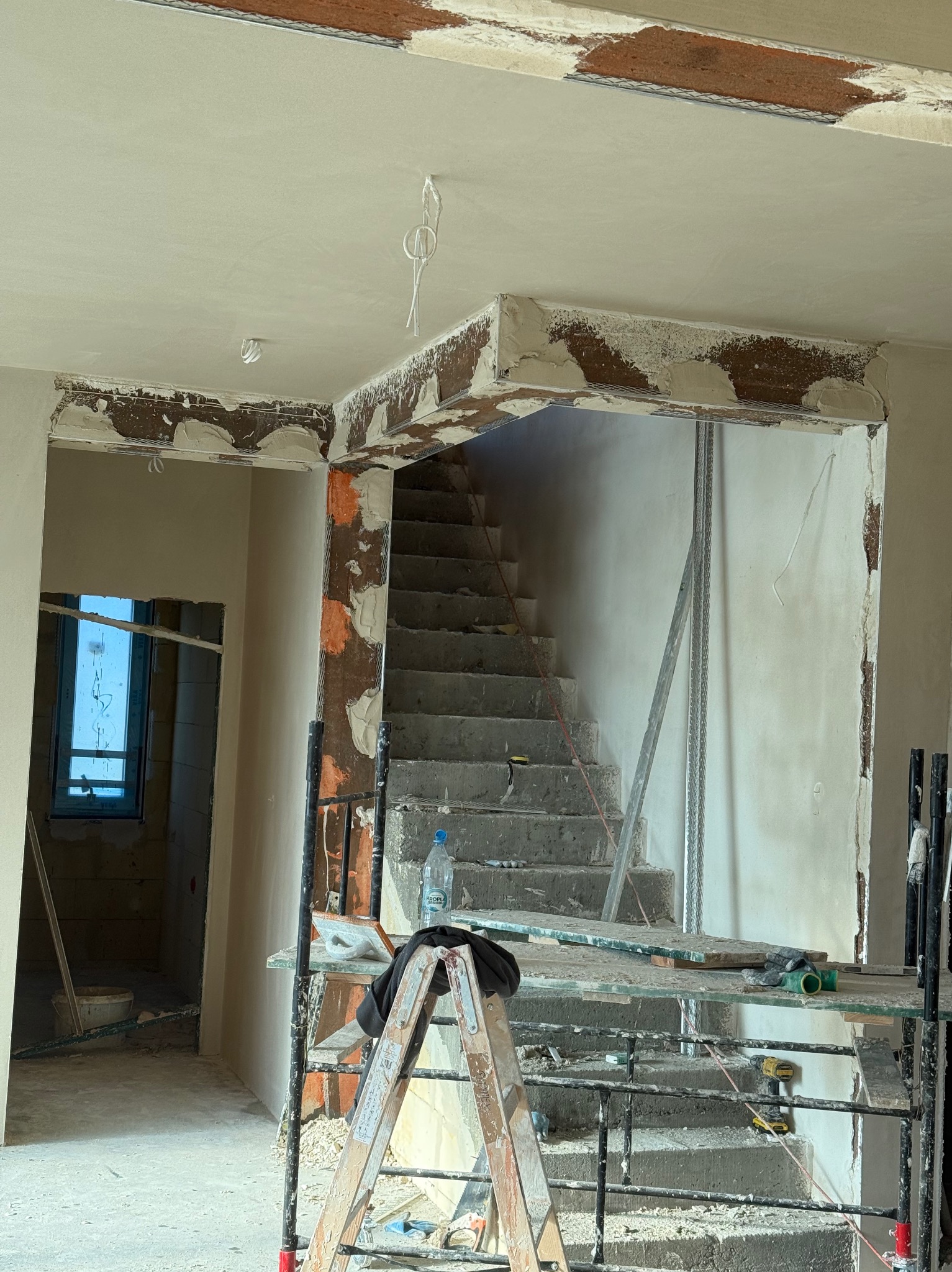 Wnętrze w trakcie prac tynkarskich: widoczne schody, rusztowanie z narzędziami, fragment okna i ściany z nałożonym tynkiem w surowym stanie.