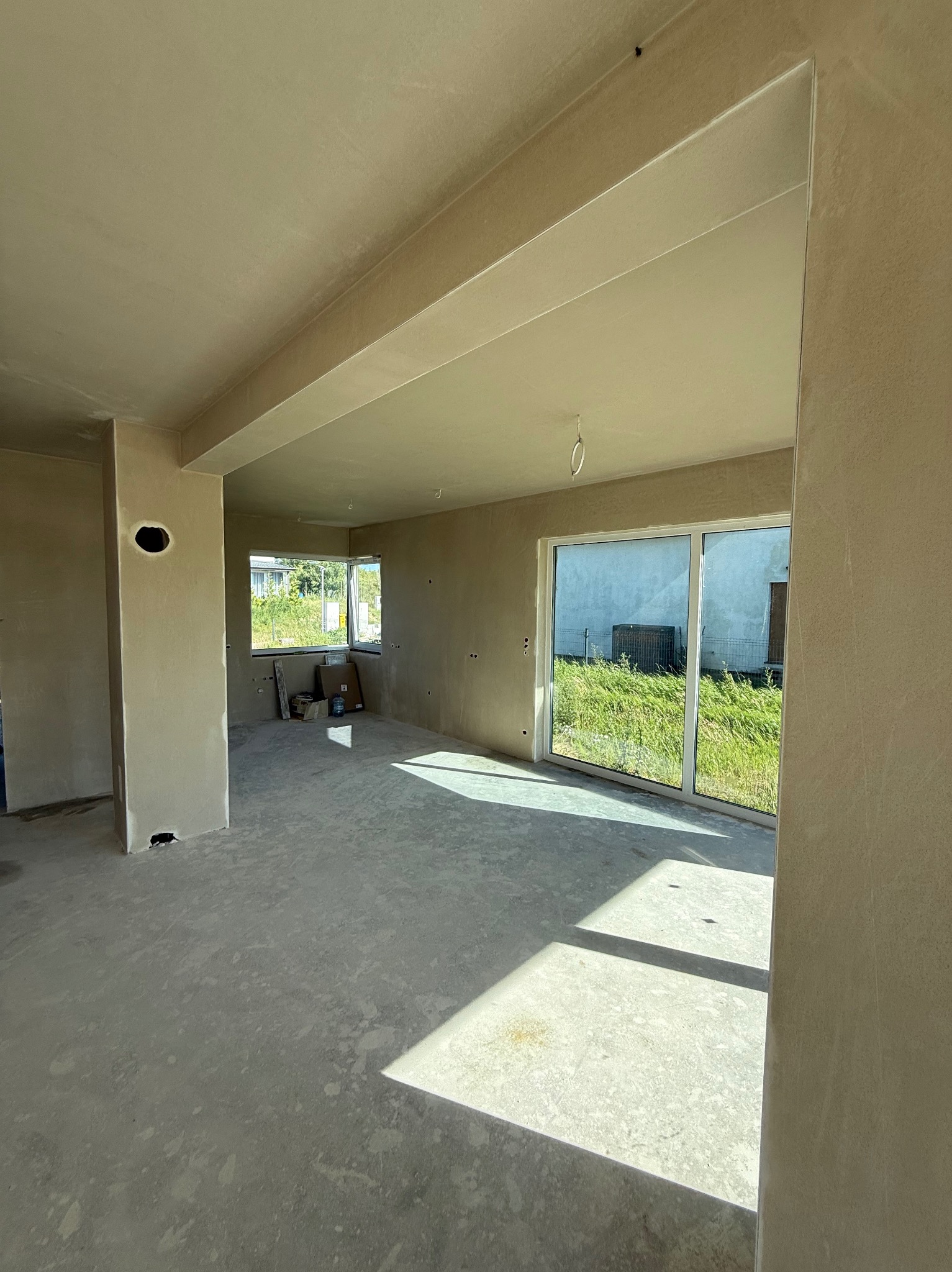 Jasne wnętrze po nałożeniu tynków; widoczne ściany, sufit, podłoga, okno z widokiem na zieleń, wpadające promienie słoneczne. Nowoczesne wykończenie.