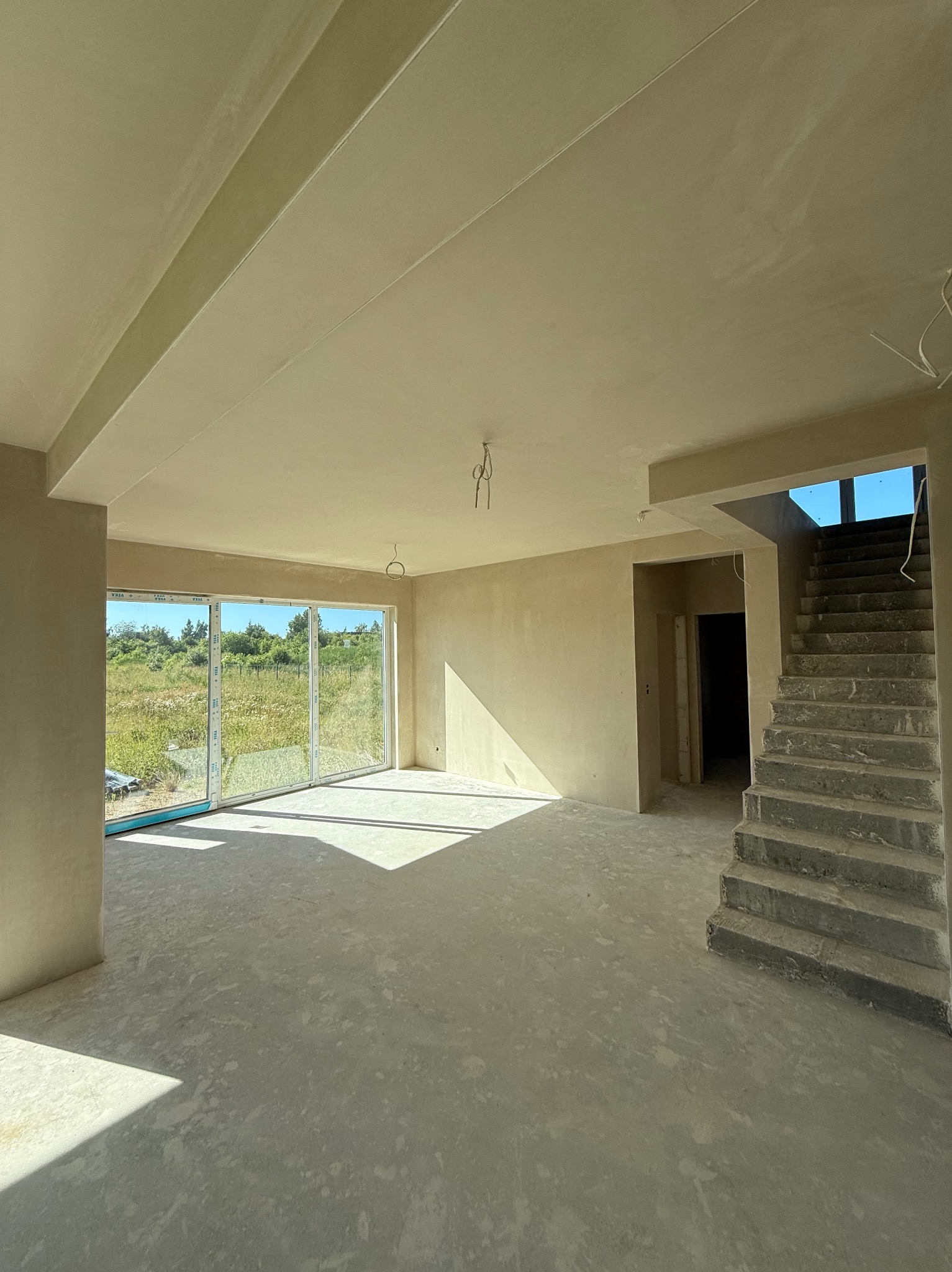 Wnętrze domu po nałożeniu tynków: jasne ściany, surowa podłoga, schody w tle. Duże okno z widokiem na zieleń. Delikatne światło podkreśla gładkość powierzchni.